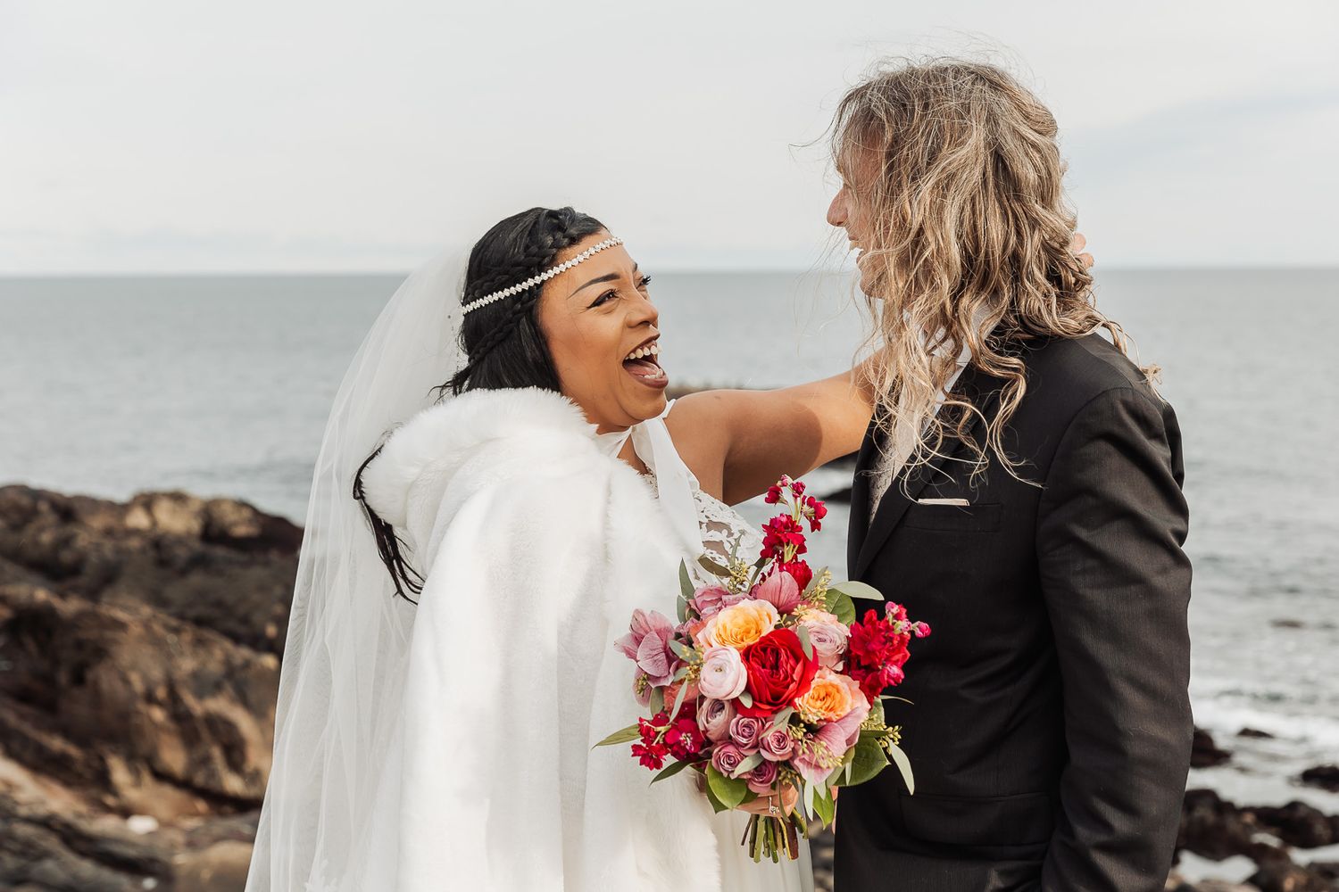 A joyful moment between newlyweds holding pink and red bouquet against rocky ocean coastline on their wedding day.