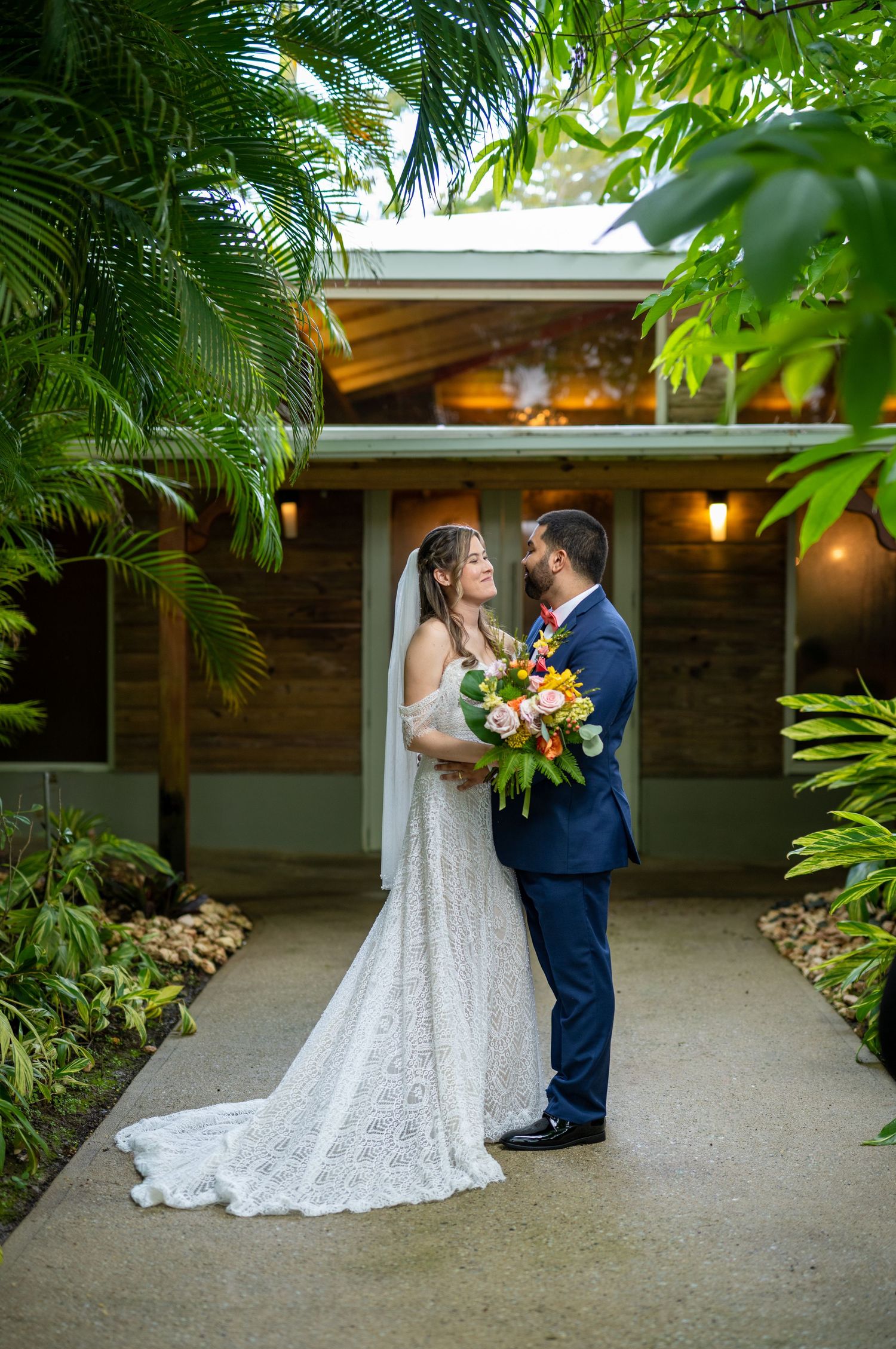 Newlywed couple shares romantic moment on garden pathway surrounded by lush tropical palm fronds and greenery. Zafra del Caribe Gurabo