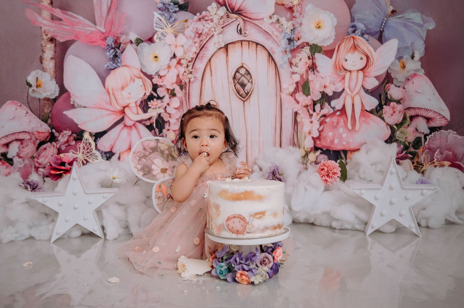 Elegant first birthday setup with fairy door backdrop, pink florals, stars and white tulle decorations surrounding cake.