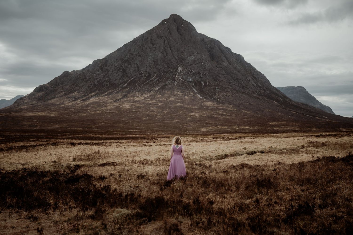 A lone figure in a purple dress stands in the barren Scottish Highlands beneath a dramatic mountain peak and moody sky.
