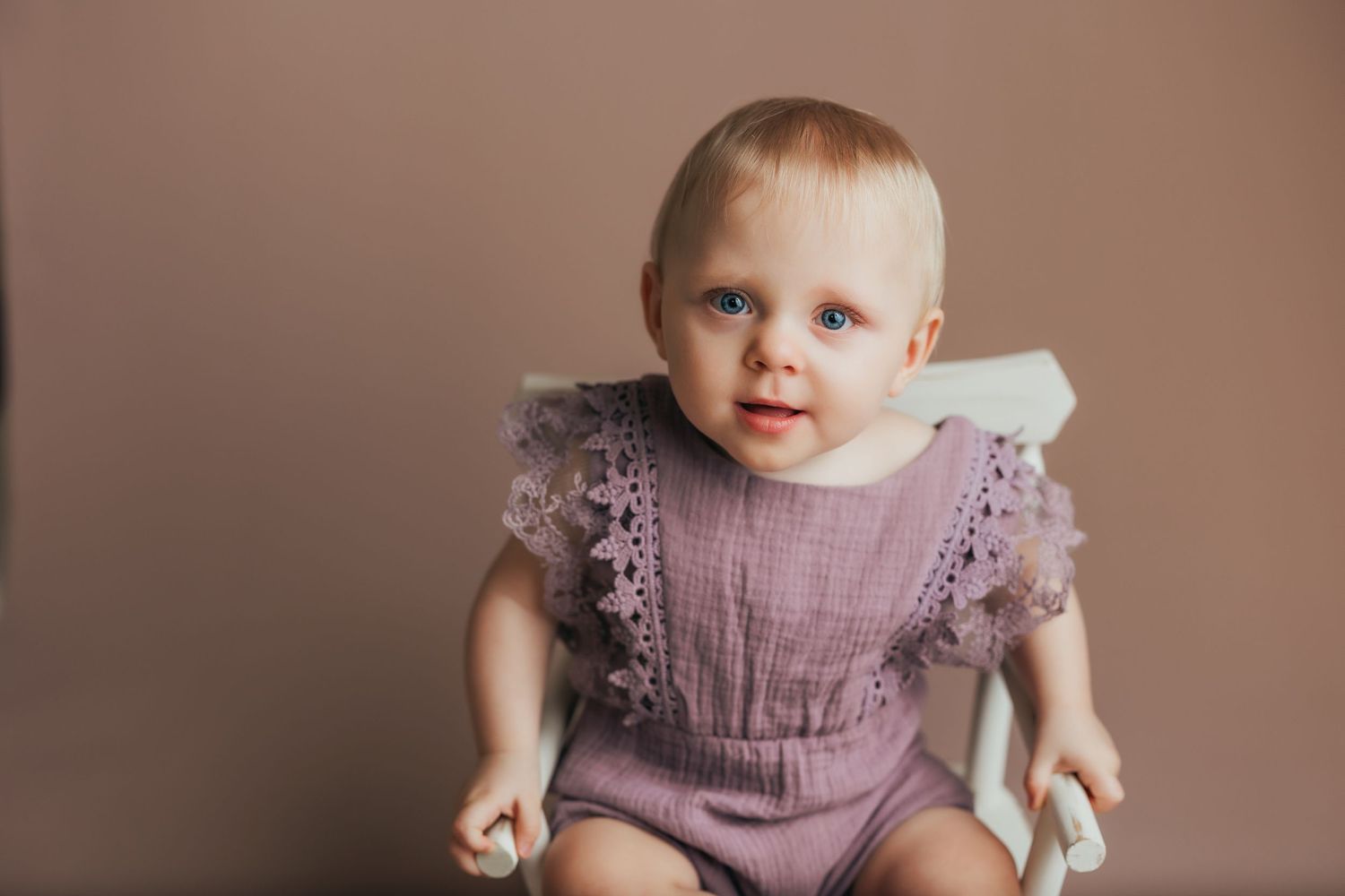 Baby wearing mauve lace-trimmed romper poses during studio photography session.