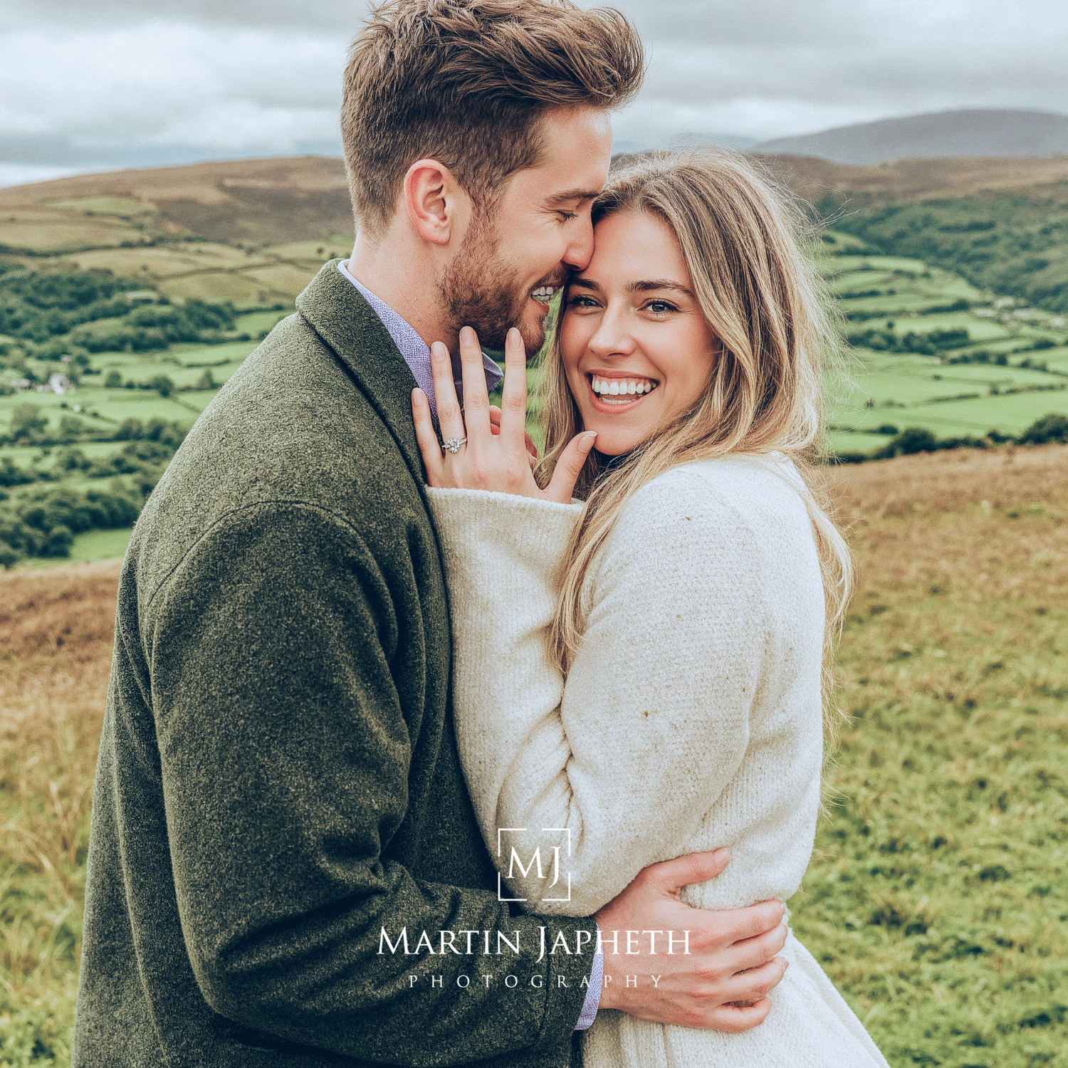 Happy couple in sweaters embracing and showing engagement ring against scenic green rolling hills landscape.