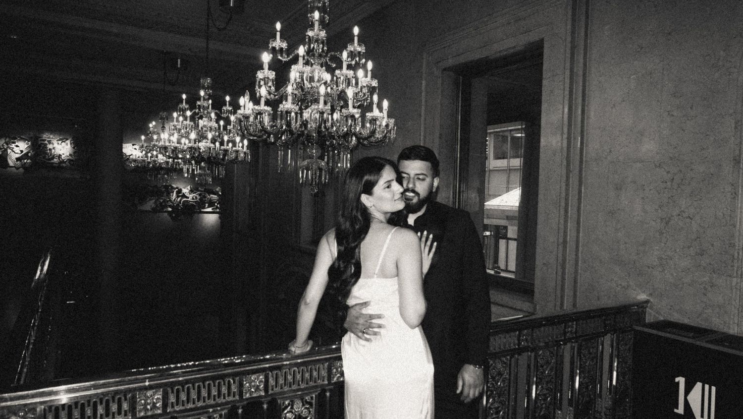 A romantic couple shares an intimate moment on a balcony under elegant crystal chandeliers in this vintage black and white photo.