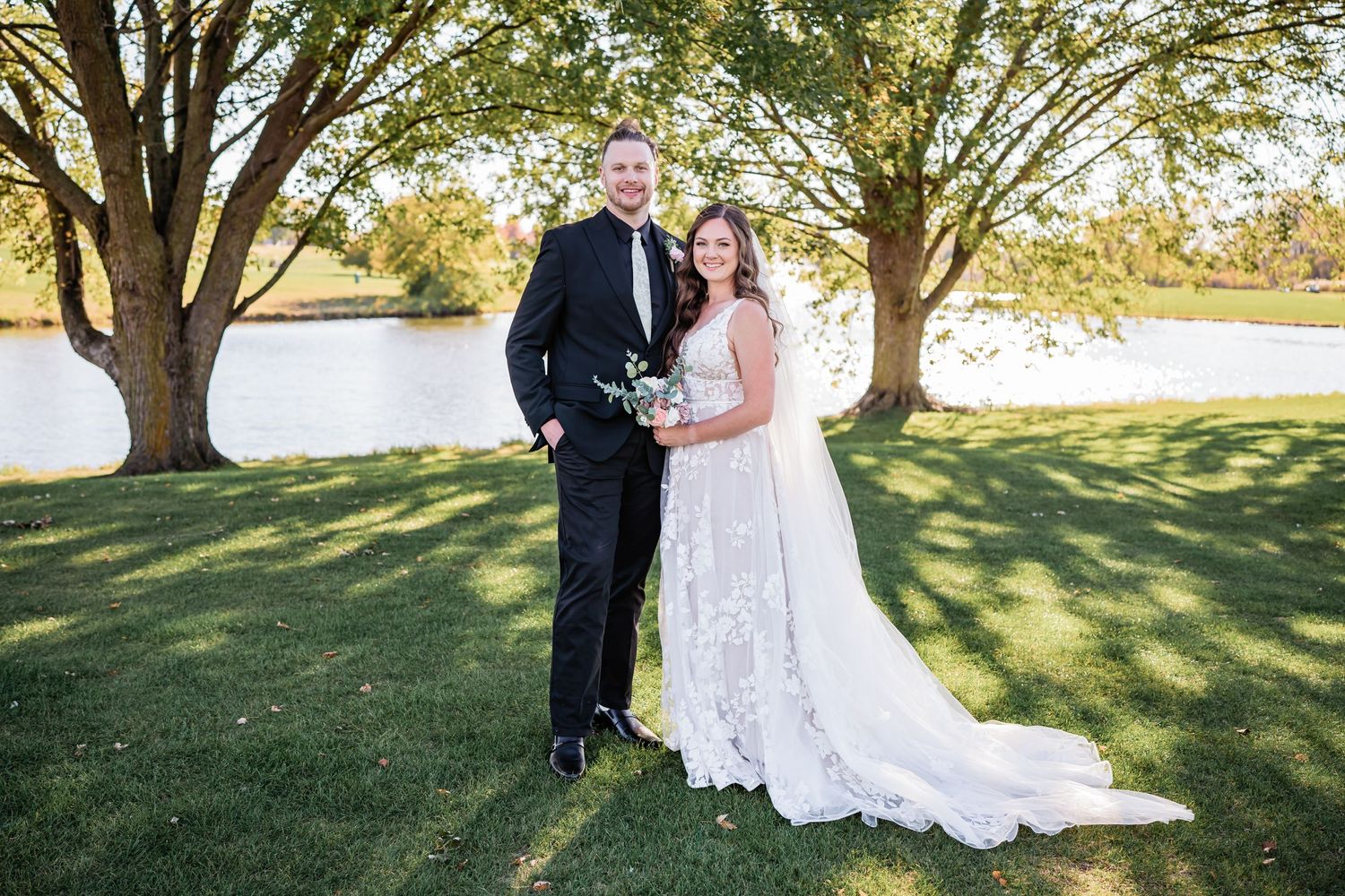 A couple in formal wedding attire poses under large trees by a lake on a sunny day.