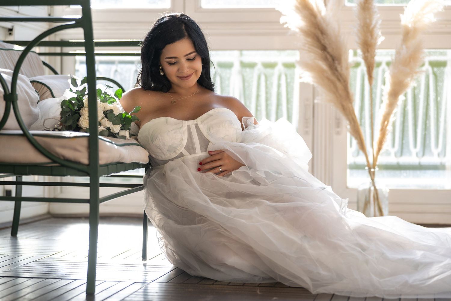A person in a flowing white off-shoulder wedding gown poses gracefully on a porch with pampas grass decor.