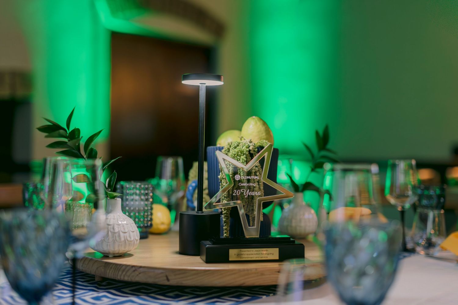 Elegant event table centerpiece with trophy, greenery, and glassware under green lighting.