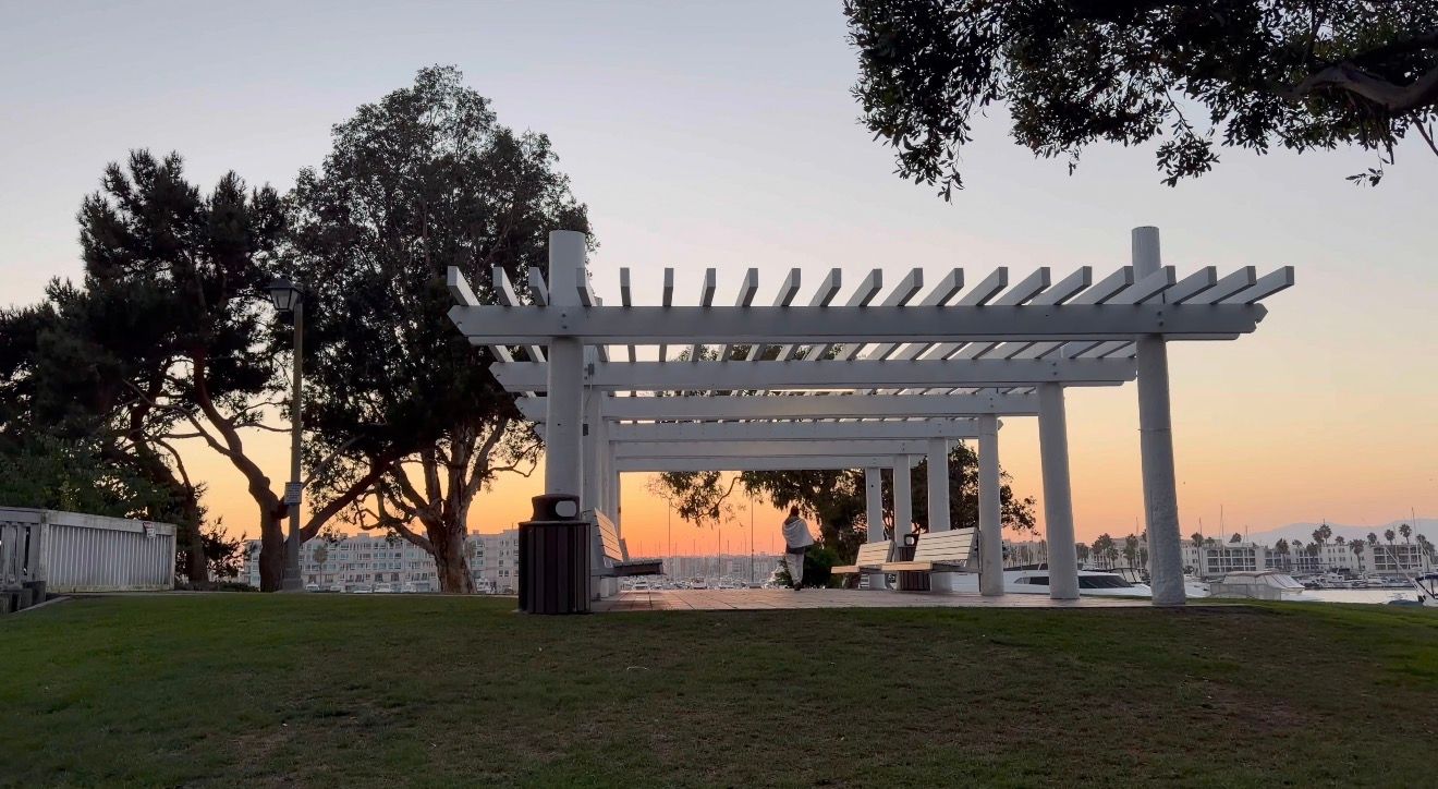 White pergola structure overlooking a sunset view from a hilltop park with trees silhouetted against the orange sky.