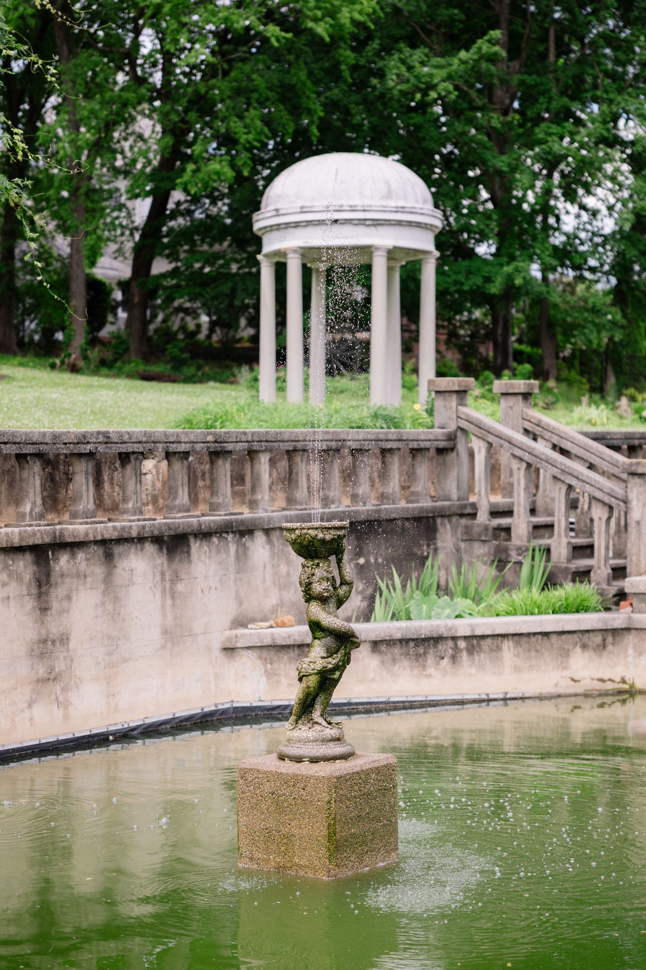 A classical white domed gazebo and stone statue overlook a tranquil pond in a lush garden setting.