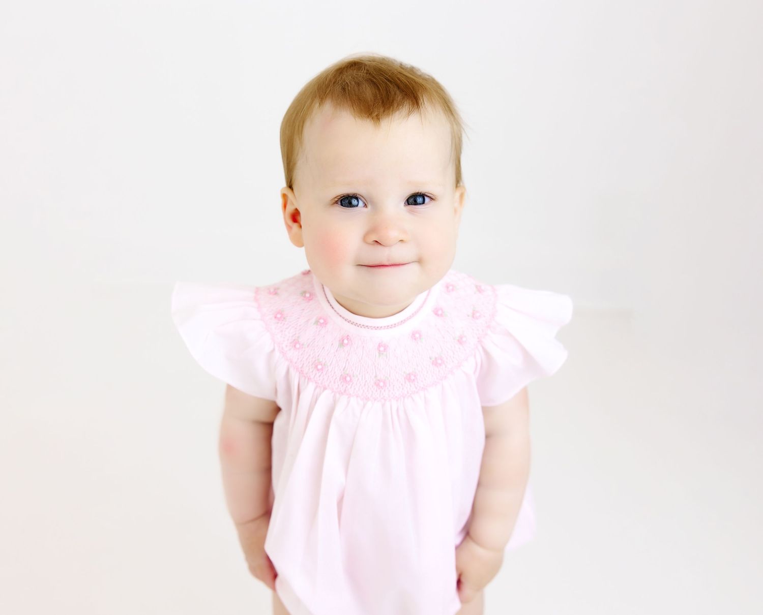 Baby wearing pink ruffled dress smiles while standing against white background.