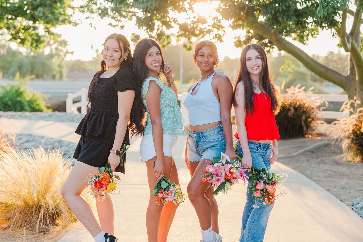 Group in casual summer outfits holding colorful flower bouquets poses together at sunset in garden setting.
