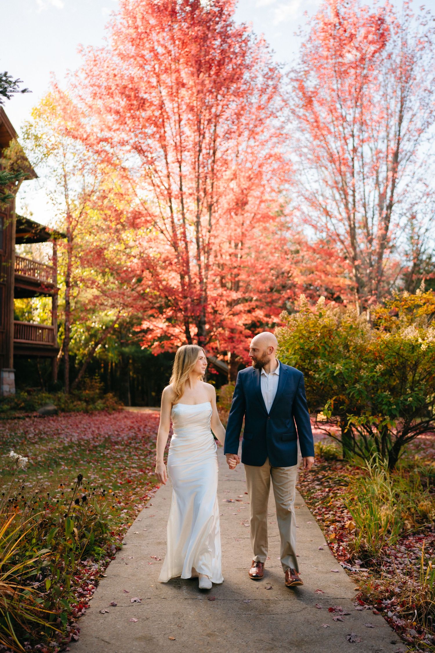 A couple walks hand in hand along a garden path surrounded by vibrant red autumn foliage on a fall day.