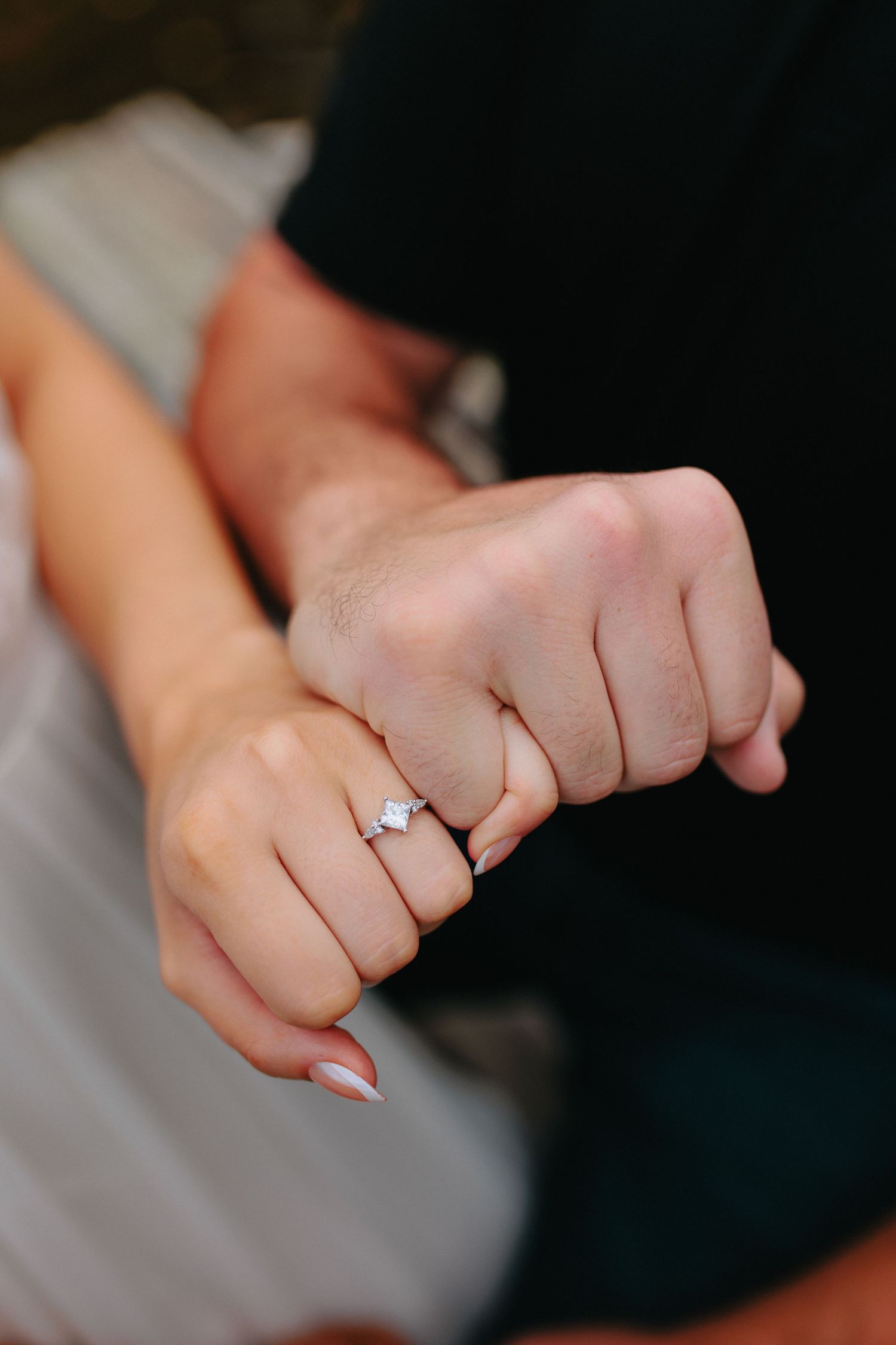 Close-up photo of hands wearing a diamond engagement ring on a dark background.