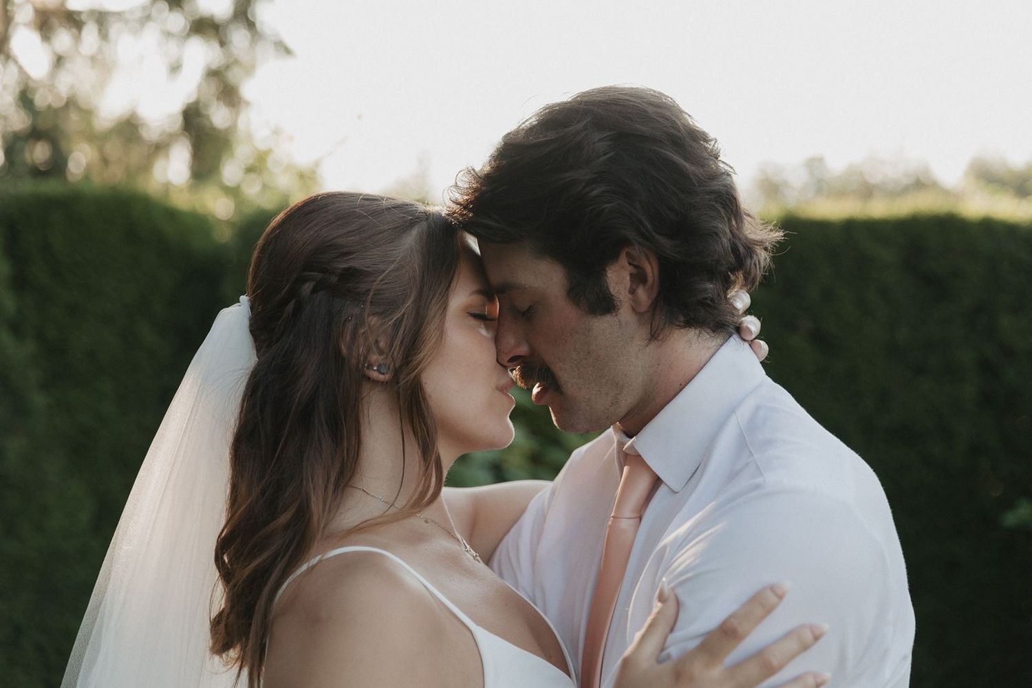 A bride and groom share an intimate forehead-to-forehead moment outdoors surrounded by lush green hedges.