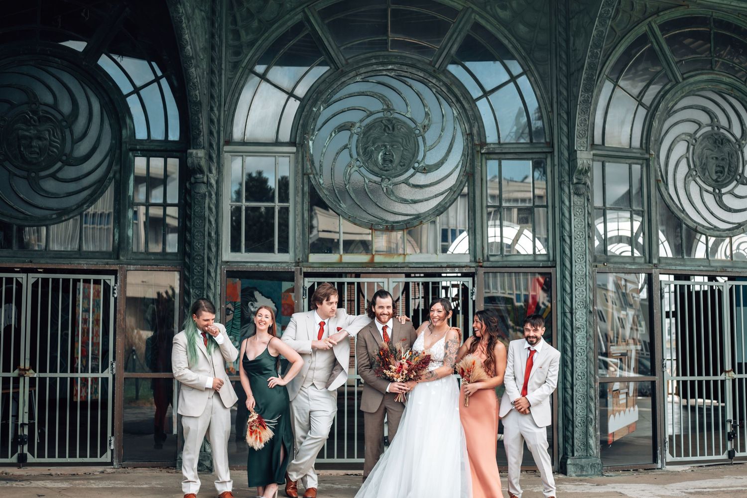 Wedding party wearing orange and white poses near elaborate glass decor at a modern venue entrance.