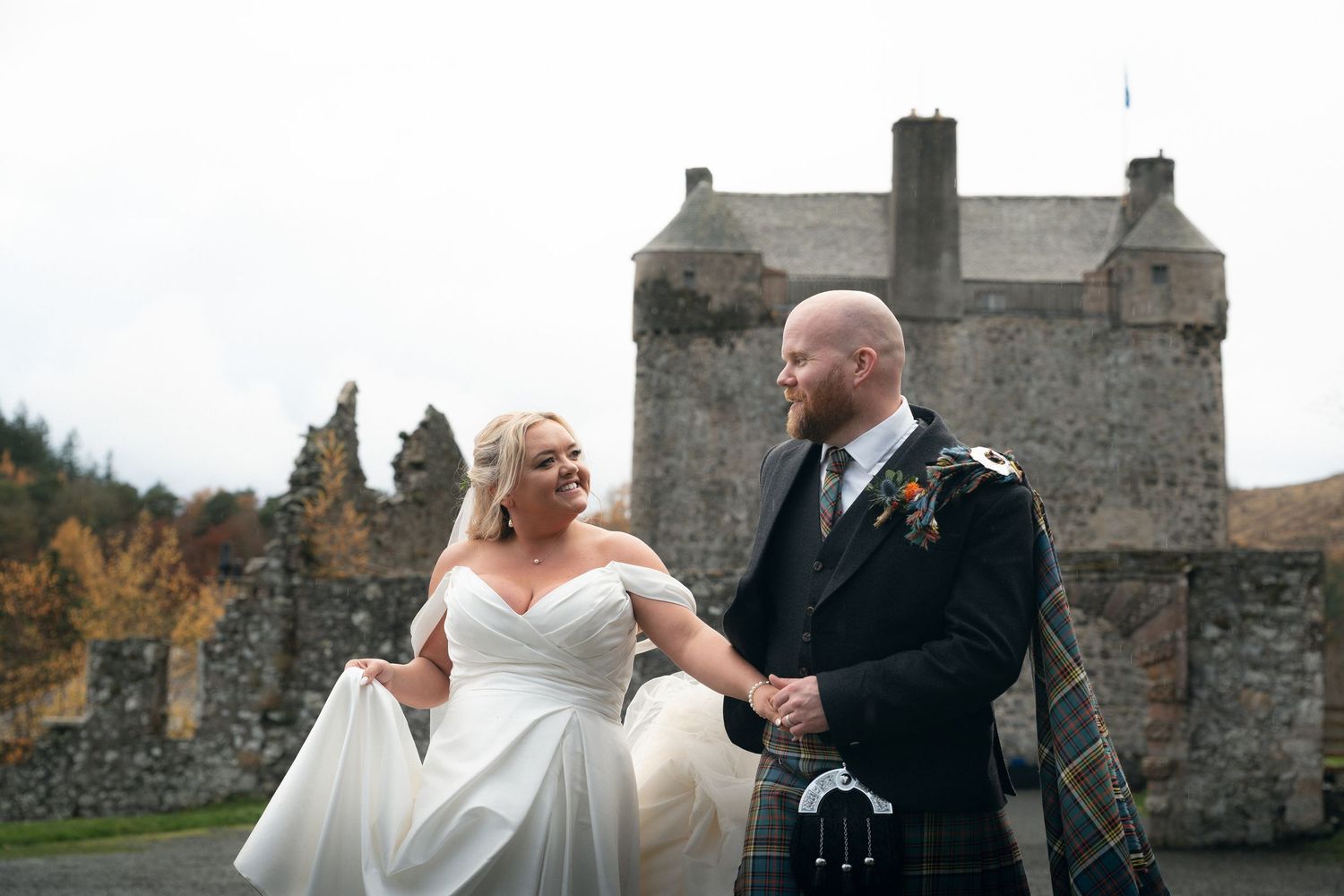 Couple walking through a garden path in wedding attire on a rainy day with umbrellas.