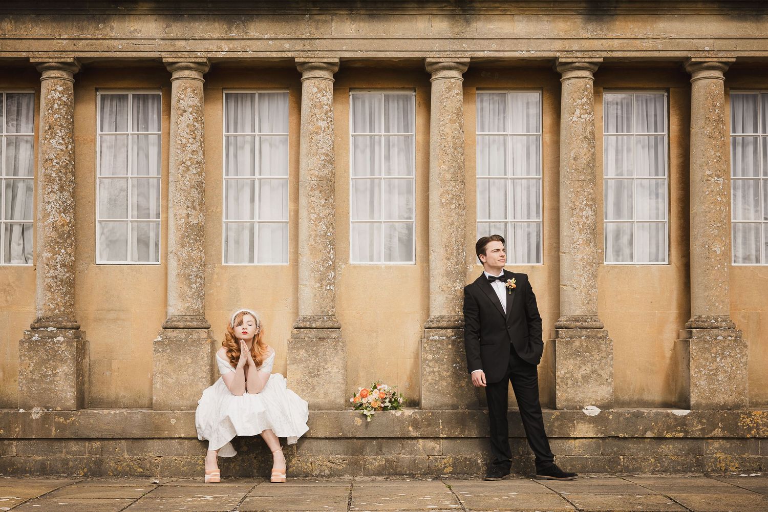 A person in a white wedding dress poses playfully on stacked vintage wooden chiavari chairs against a rustic wall.