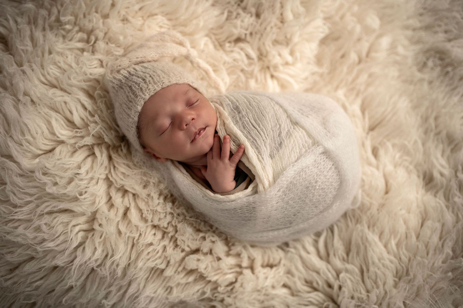 Newborn baby swaddled in white knit wrap sleeping peacefully on soft cream fur blanket.