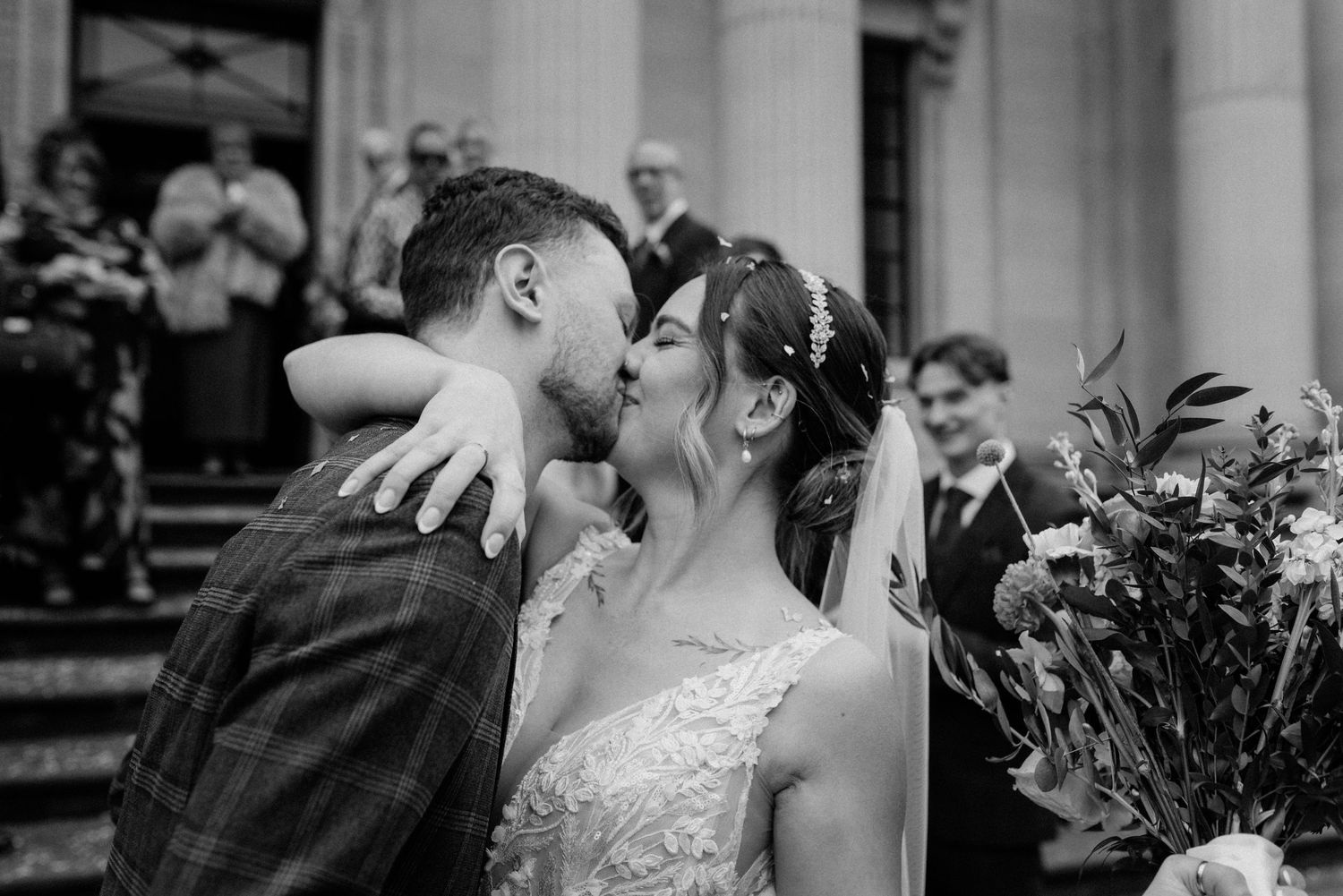 A black and white wedding photograph capturing a romantic kiss with guests and floral arrangements in the background.
