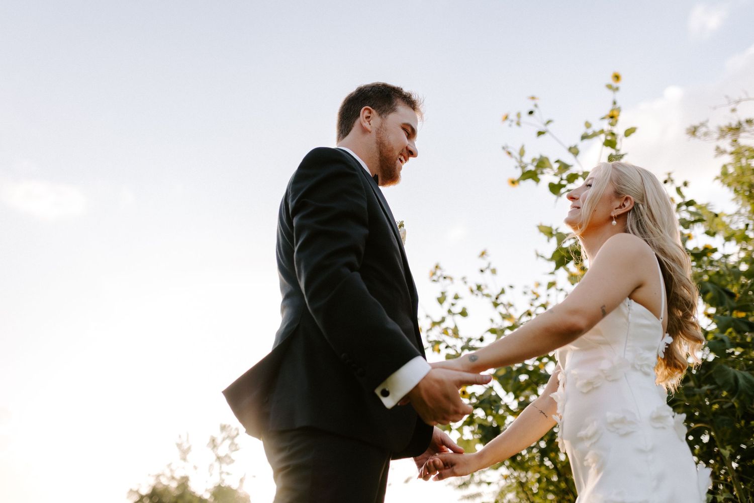 A newlywed couple poses outdoors under a large tree on their wedding day, with sunlit greenery in the background.
