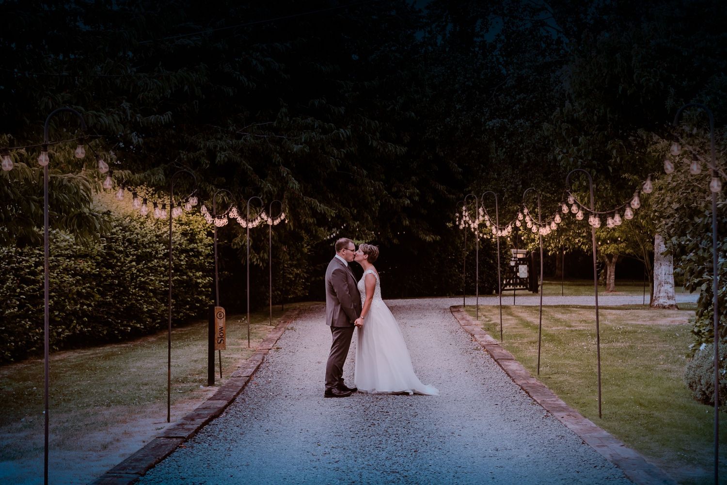 A couple embraces on a romantic path lit by twinkling string lights at dusk.