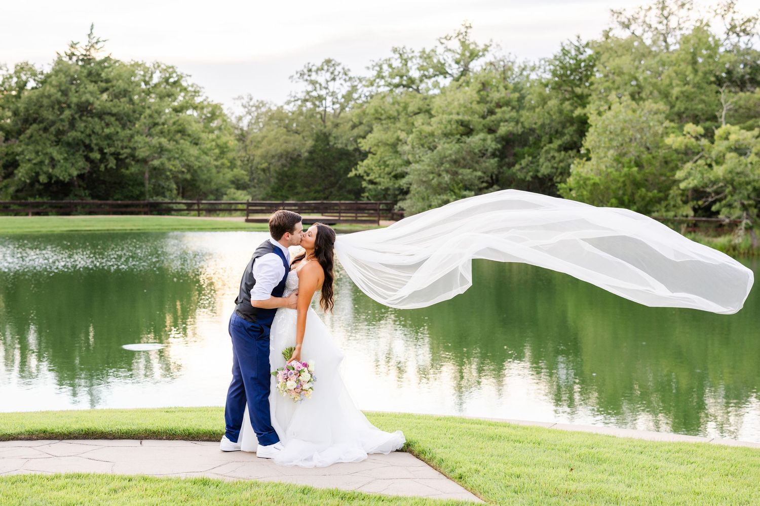 A couple in wedding attire poses by a lake while a long veil flows dramatically in the wind creating a romantic scene.