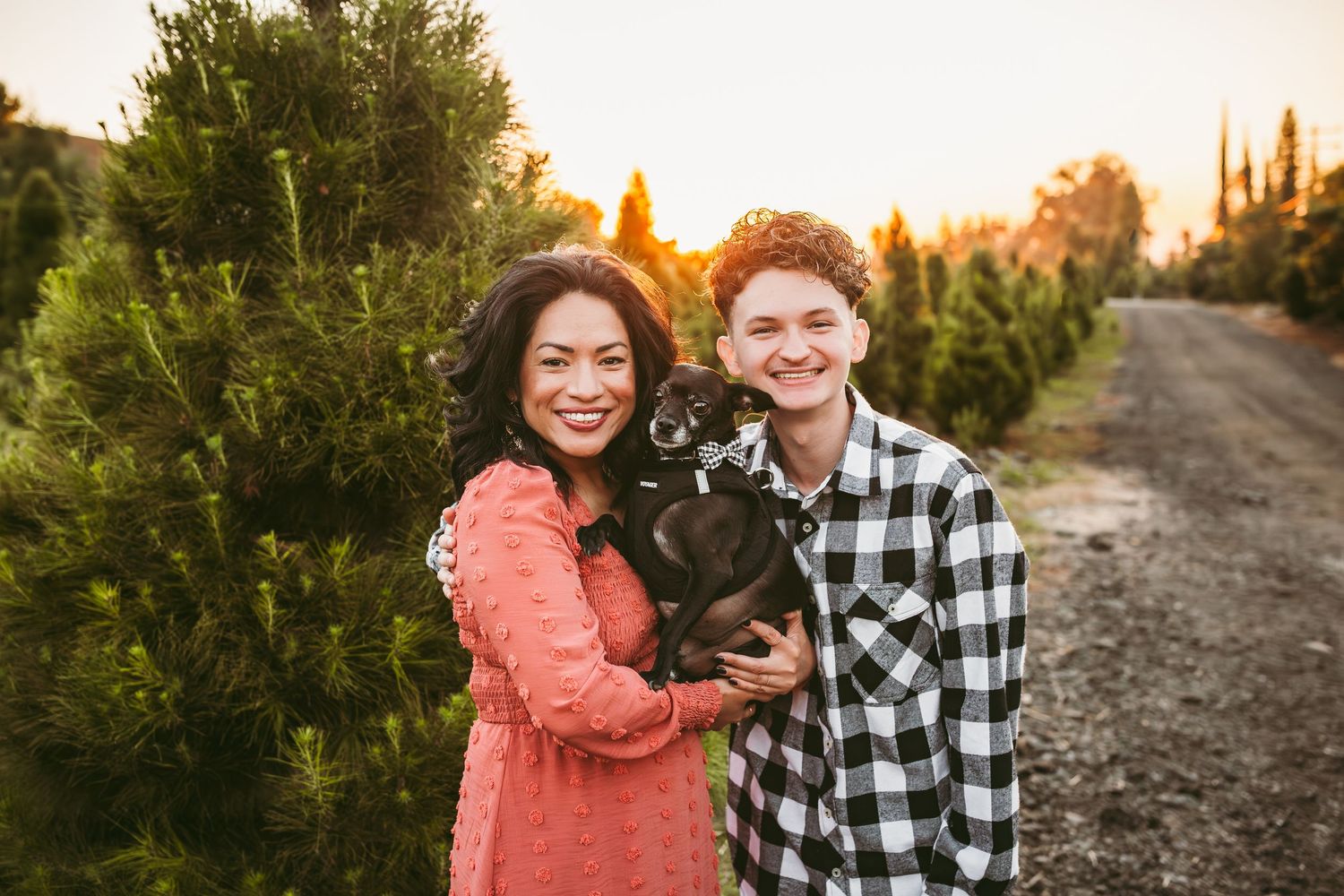 Two people holding a black dog while standing next to evergreen trees at sunset on a dirt path.