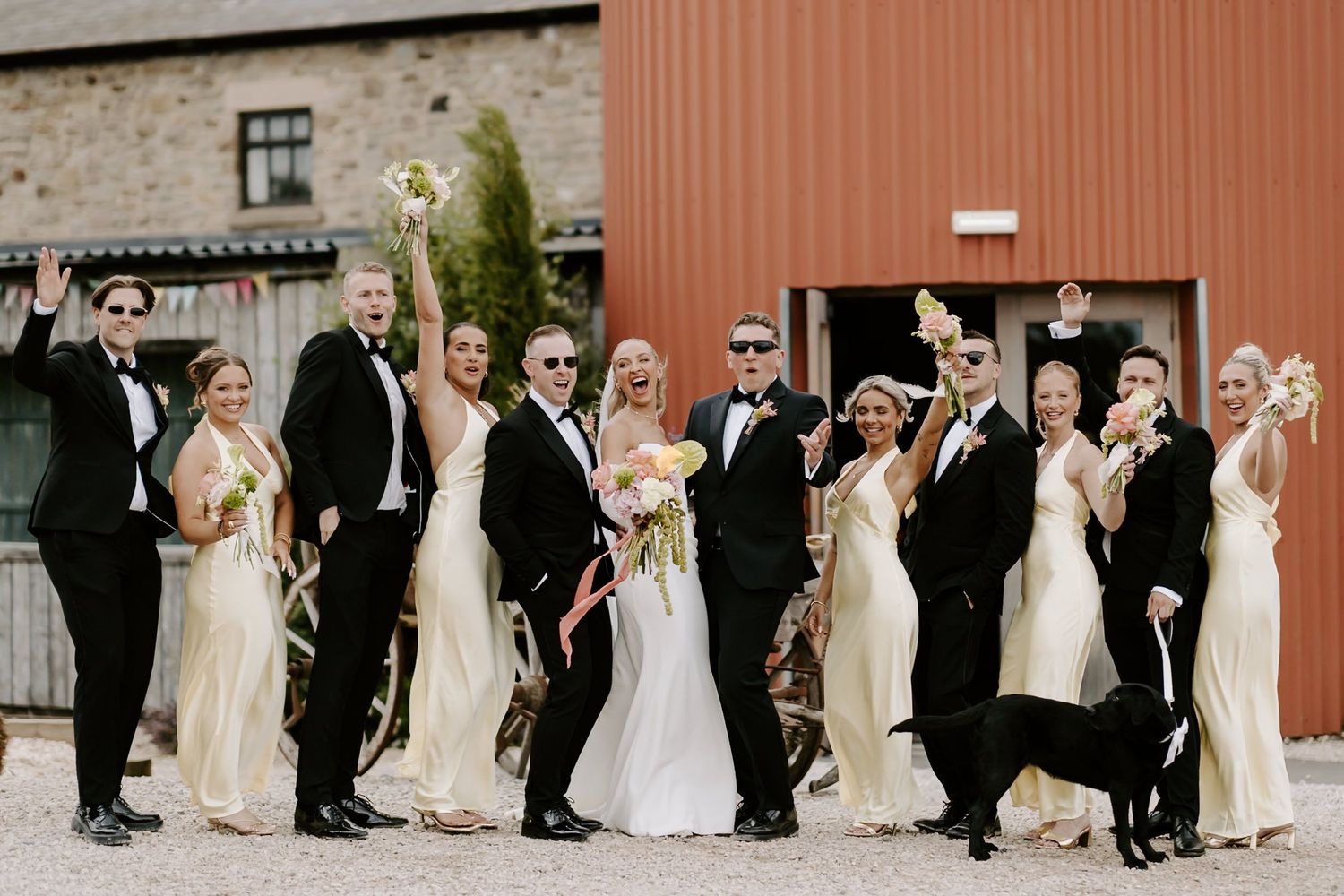 Wedding party in formal attire poses in a line against rustic red barn and stone wall backdrop.