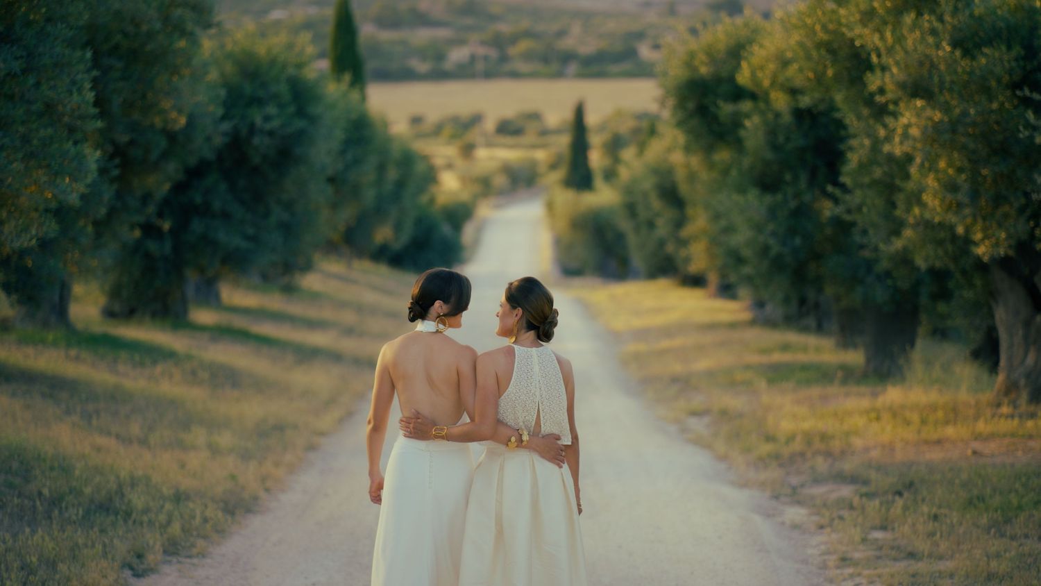 Two people walking down a serene tree-lined country road in Tuscany at sunset.