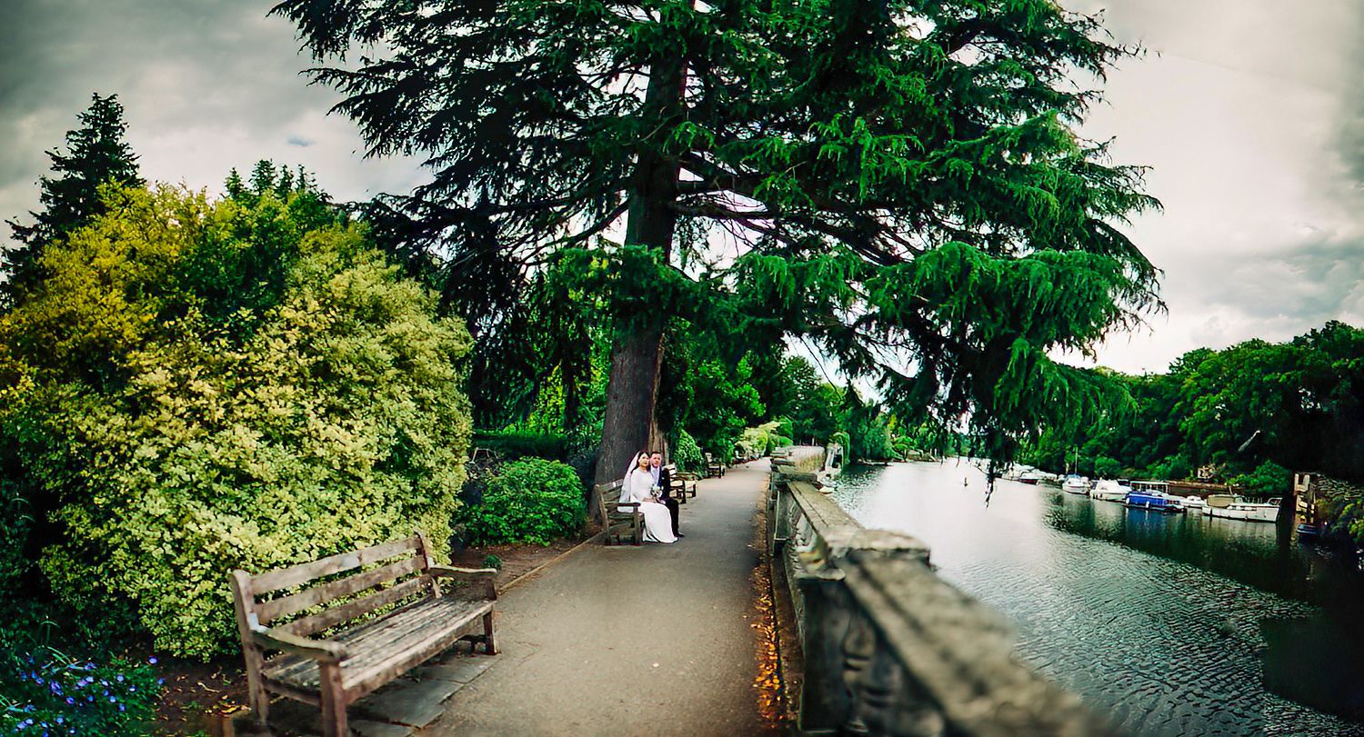 A winding path through lush gardens with wooden benches and stone railings leads to a scenic river overlook on a cloudy day.