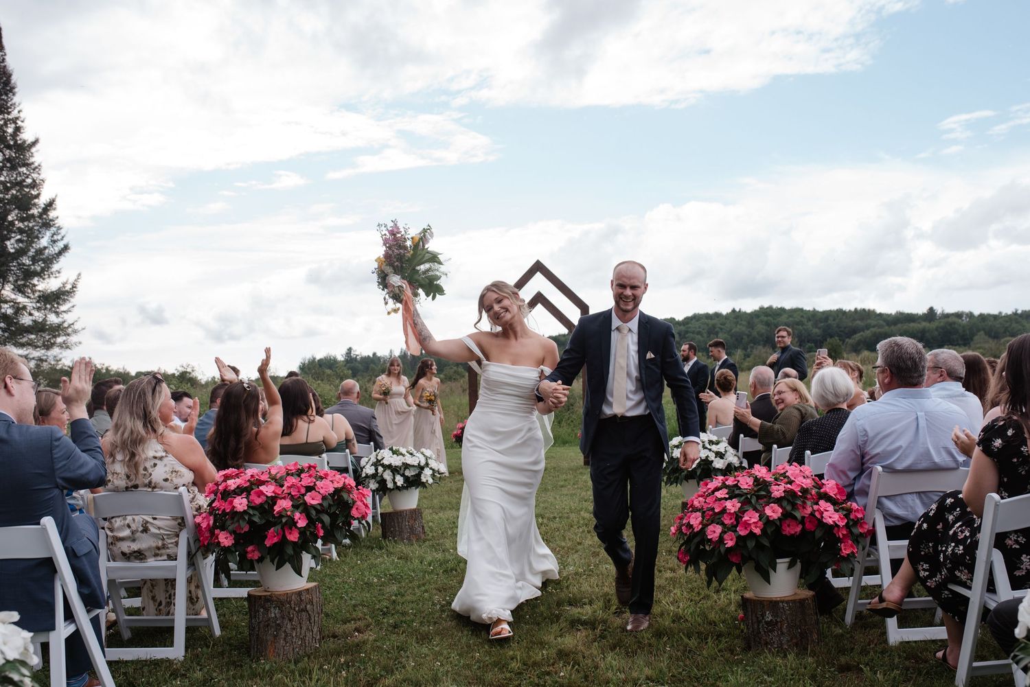 Joyful guests celebrate at an outdoor wedding ceremony with pink floral decorations and white chairs against rolling hills.