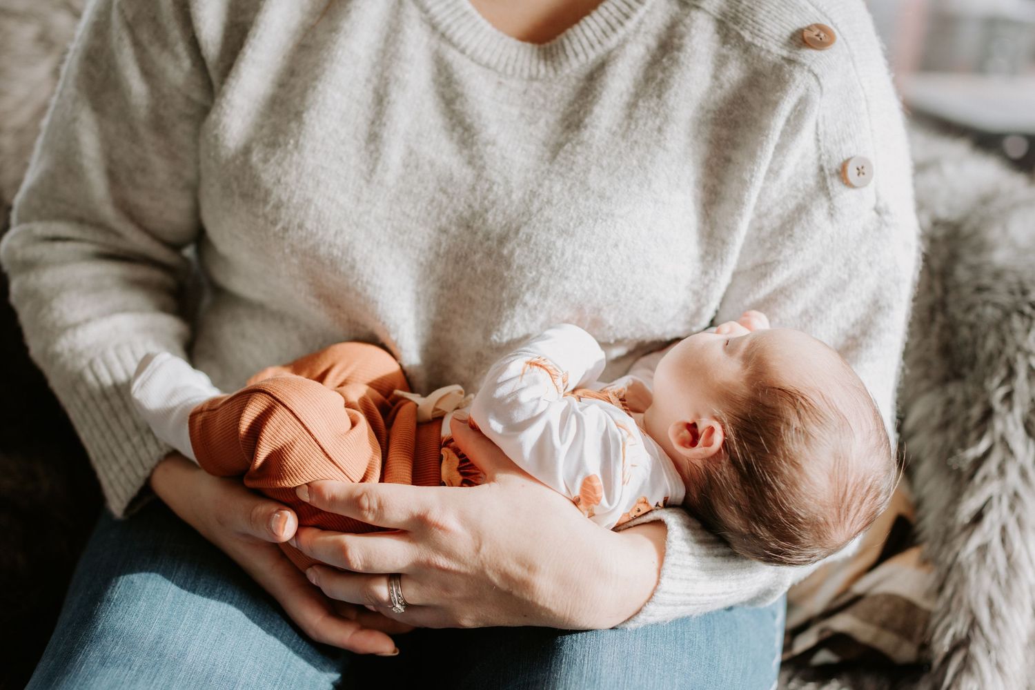 Tender moment captured as parent holds sleeping newborn baby on neutral gray bedding in soft natural light.