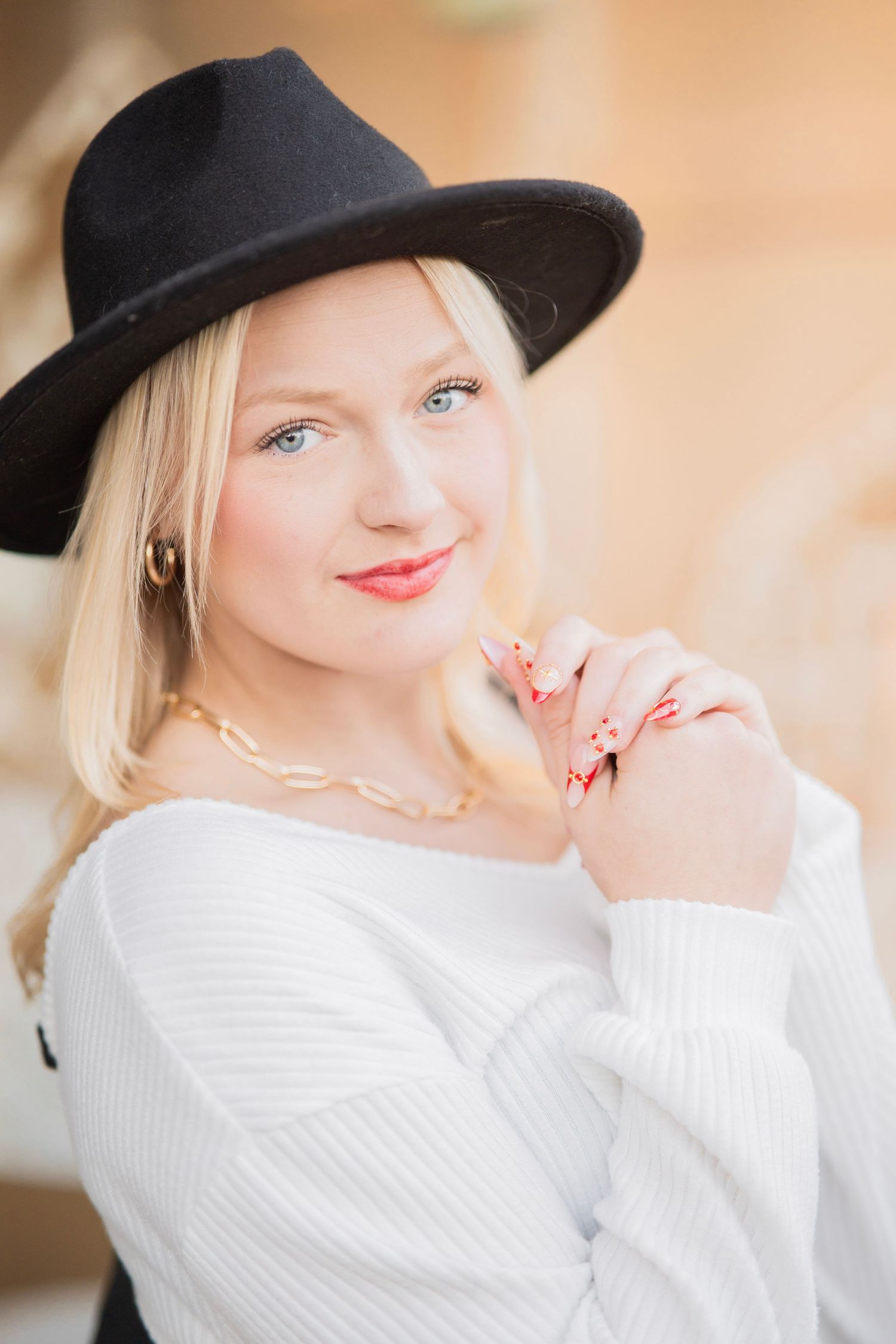 Stylish blonde woman in black fedora hat and white sweater poses with red manicured nails.