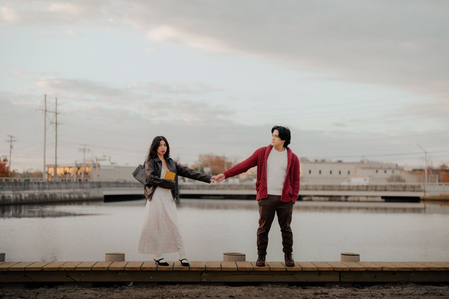 A couple holds hands at sunset on a wooden pier overlooking a peaceful harbor with industrial buildings in background.
