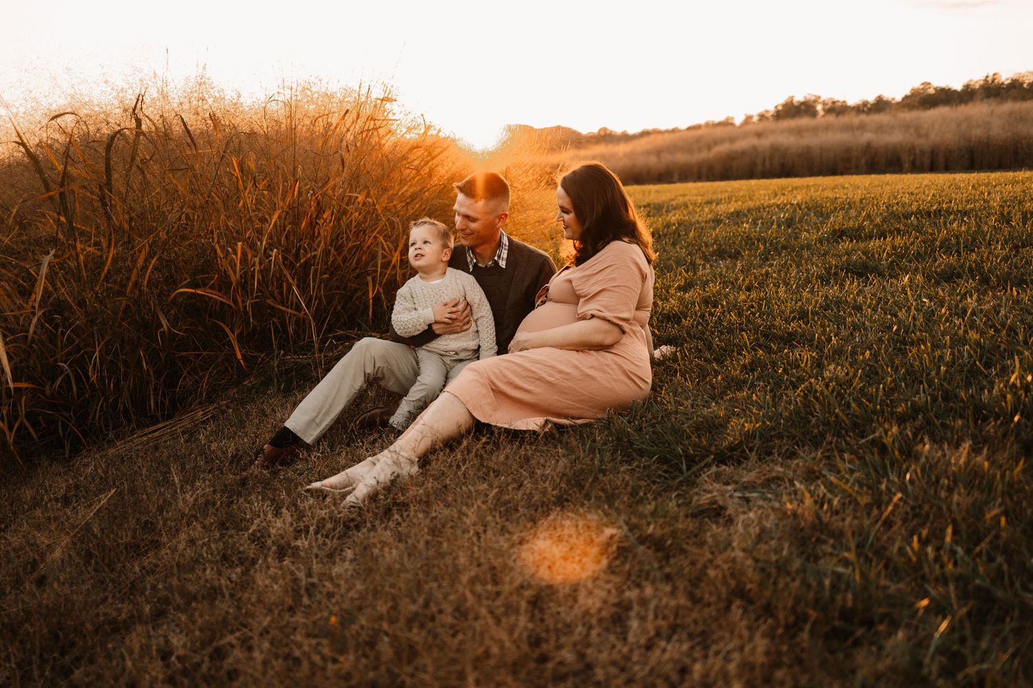 Family of three sitting in golden field at sunset, pregnant mother in pink dress with toddler and father.
