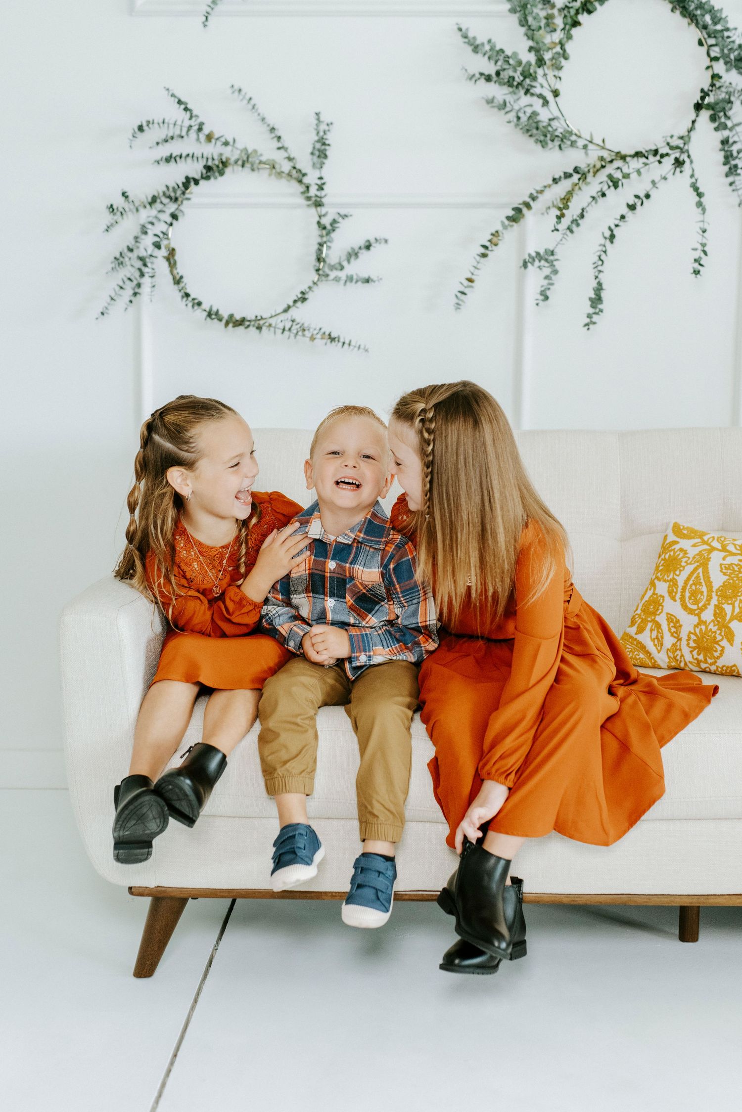 Three small children wearing orange and plaid outfits play together on a white couch under greenery decor.