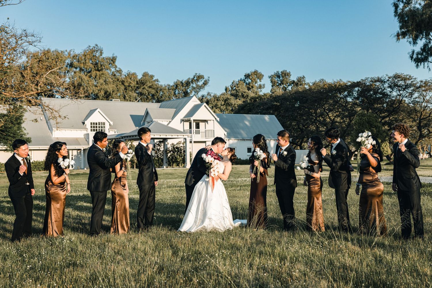 Wedding party in brown dresses stands in a line outdoors against a scenic natural backdrop on a sunny day.