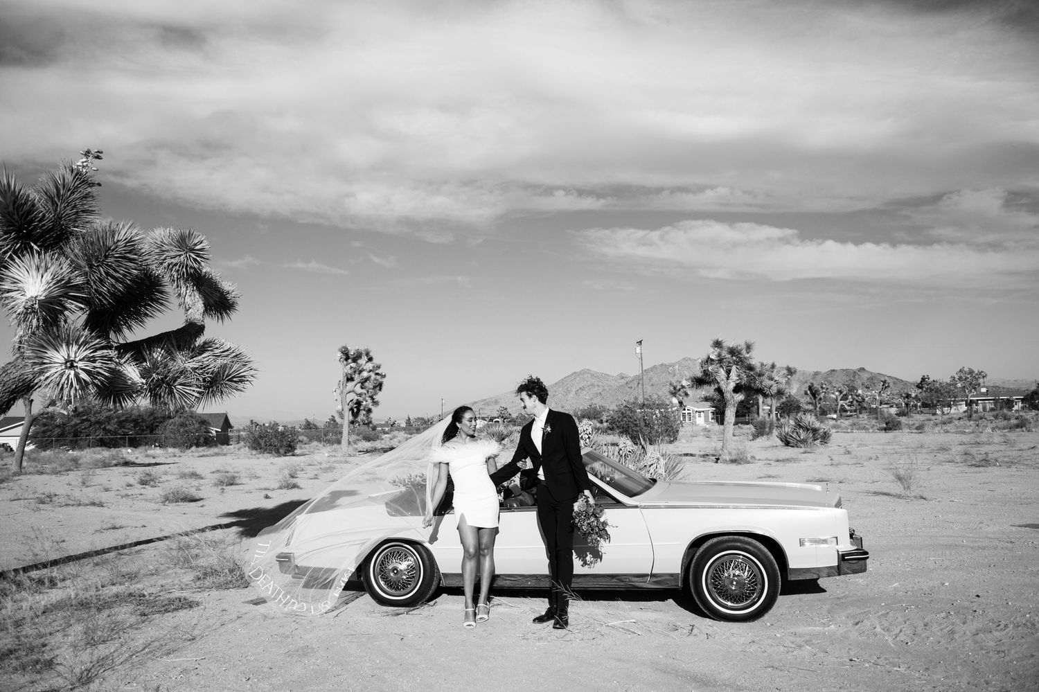A vintage white Mercedes car parked in the desert landscape with Joshua trees and two figures in formal attire.