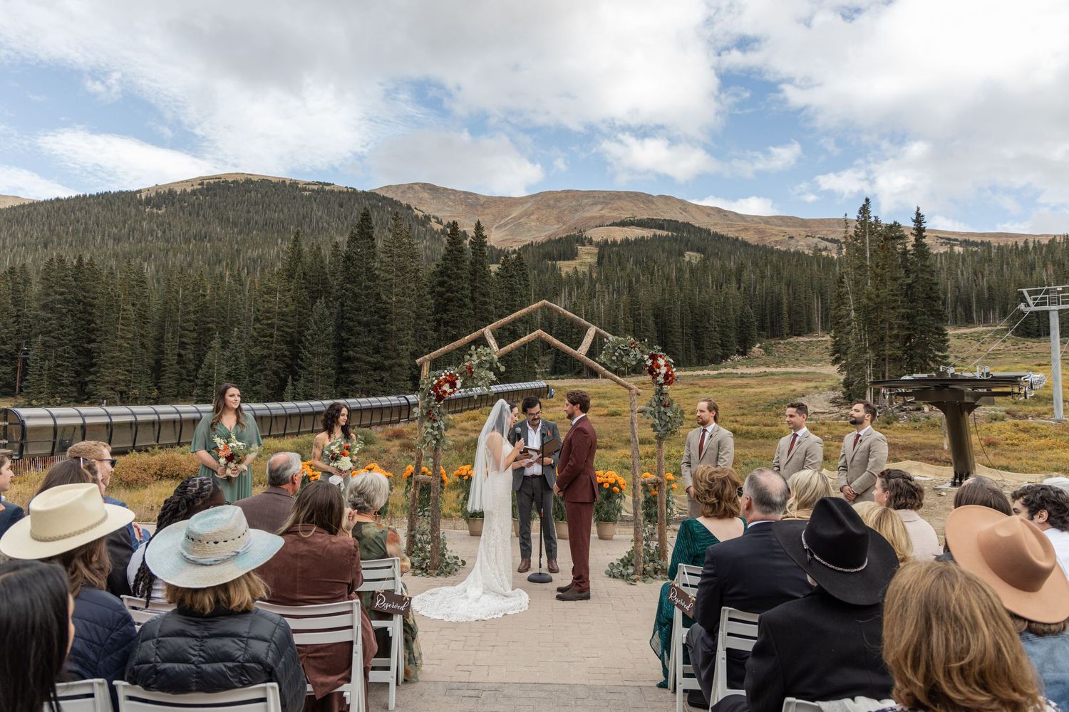 A scenic mountain wedding ceremony with guests seated outdoors surrounded by evergreen trees and mountain peaks.