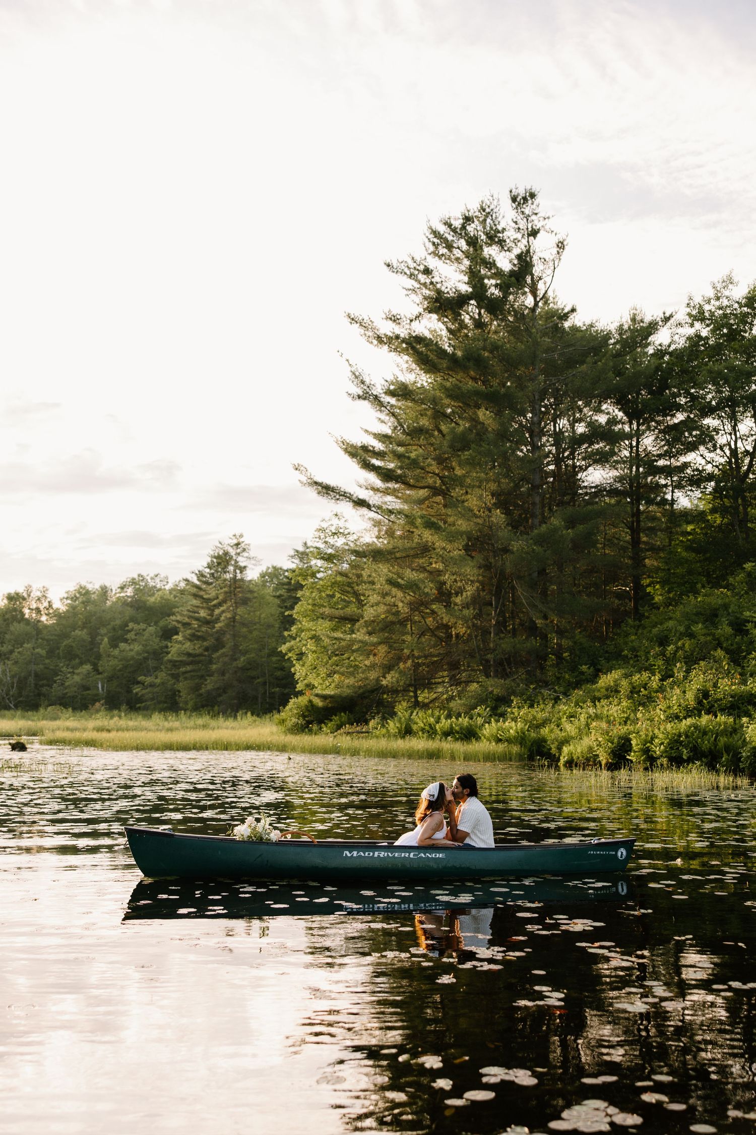 A green canoe floats on a serene lake surrounded by pine trees at sunset with lily pads dotting the water's surface.