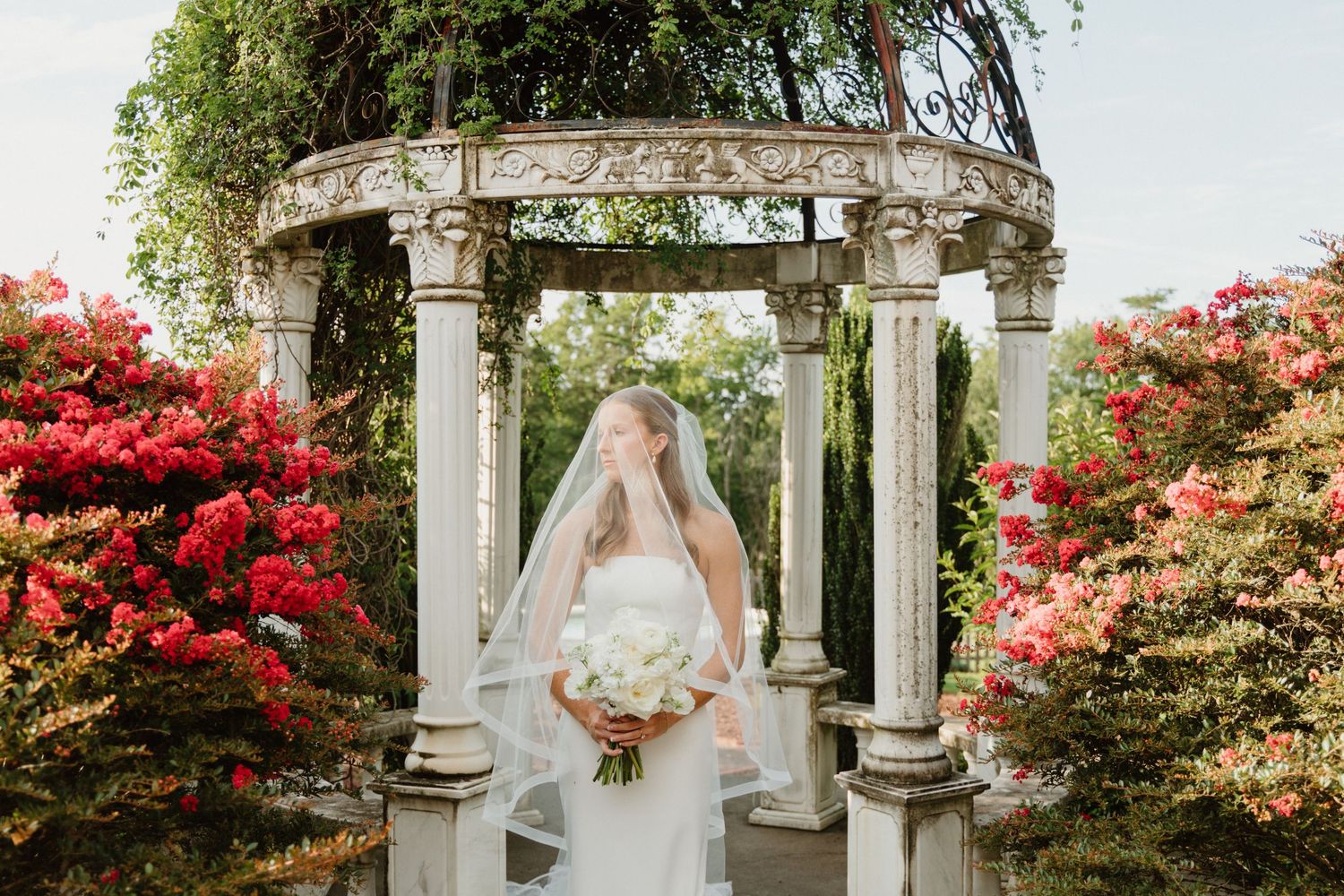 A bride in a white wedding dress stands under a stone gazebo surrounded by vibrant red autumn foliage.