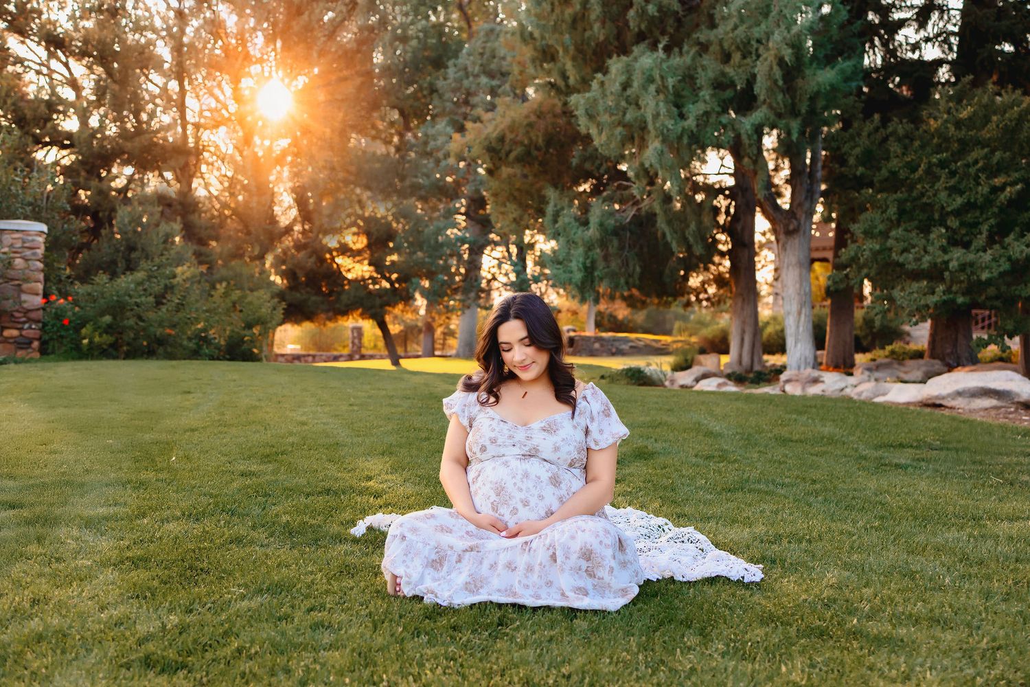 Pregnant woman in floral dress sitting on green grass at golden hour with sunlit trees in background.