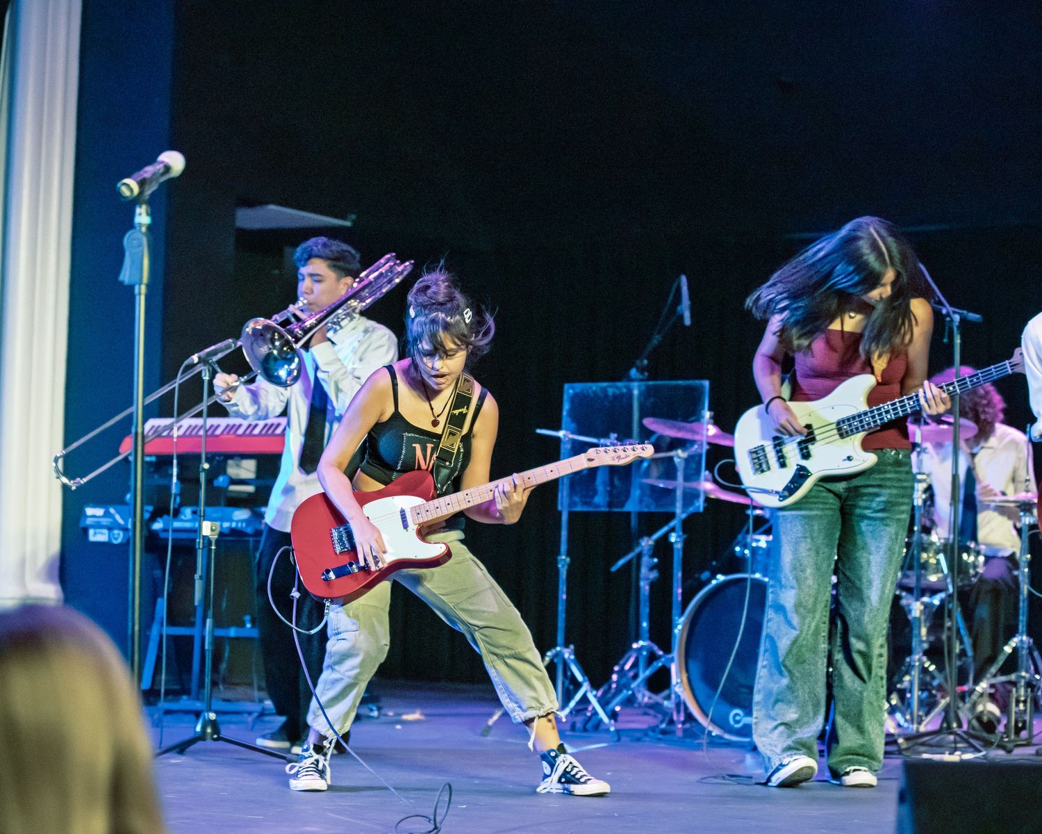 Musicians perform on stage with guitars and drums during a live concert performance with blue lighting effects.