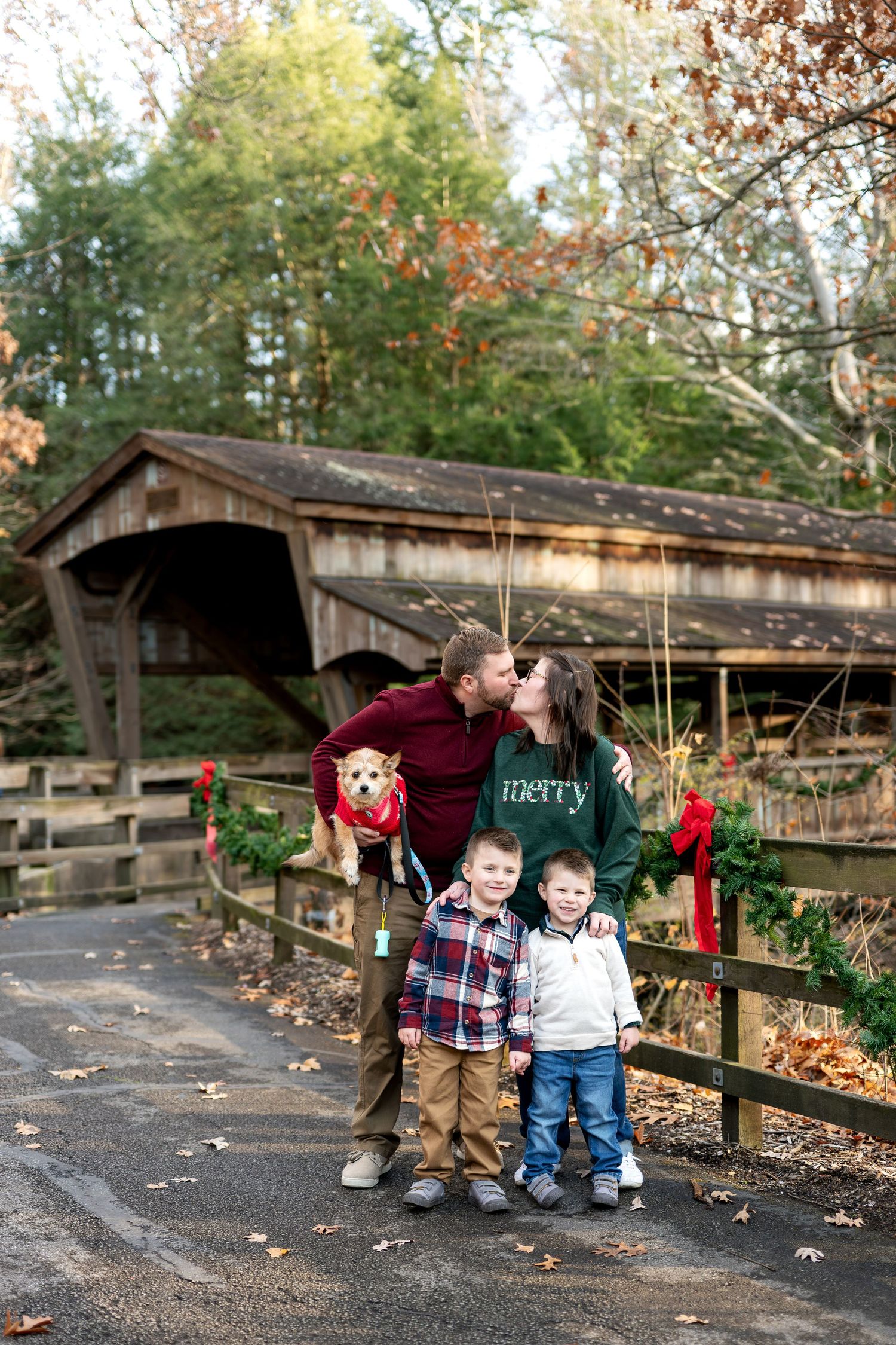 Family with two boys and a dog kissing in front of a decorated covered bridge.