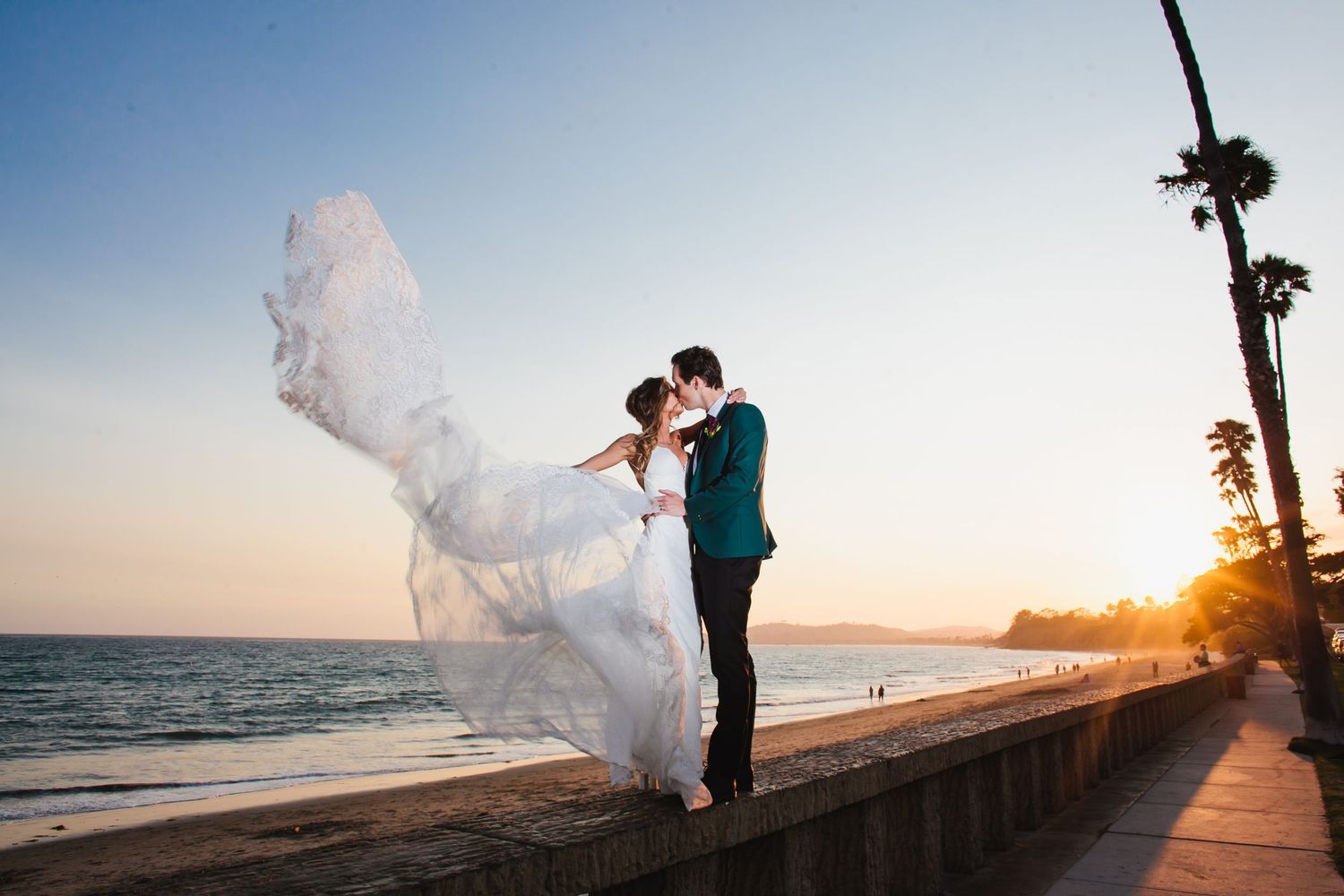 A couple in wedding attire share a romantic moment at sunset on a beachside walkway as the bride's dress flows in the wind.