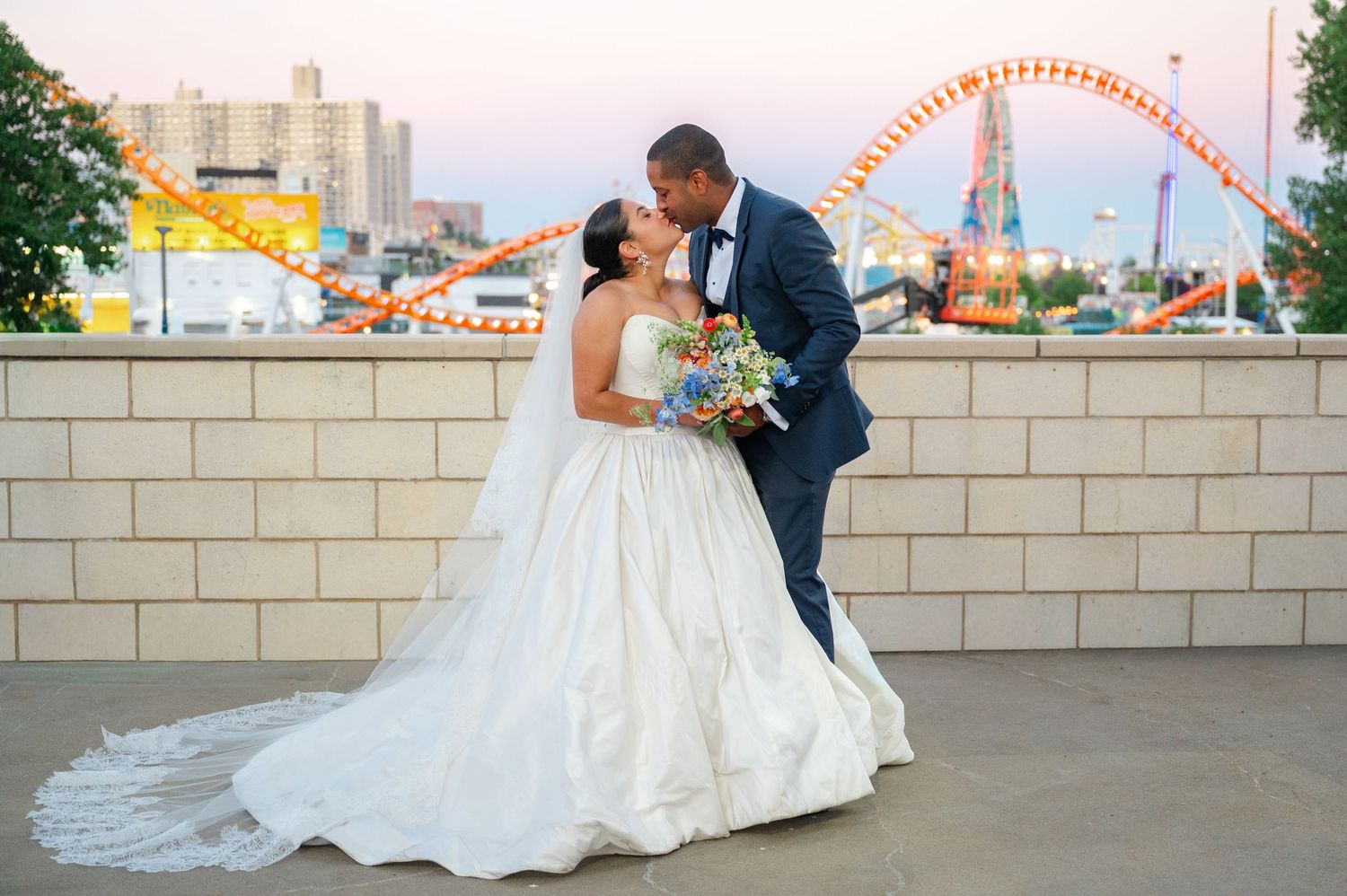 Bride in a white ball gown and groom in a navy suit kiss on with Coney Island's colorful amusement park roller coaster behind them.