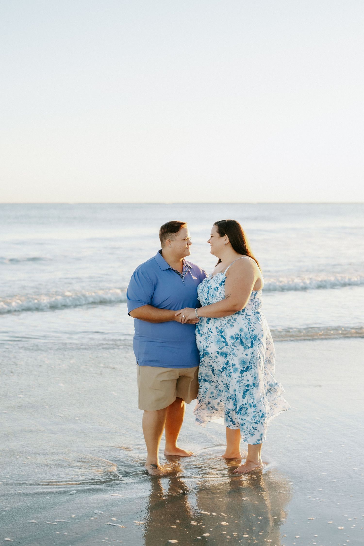 Smiling couple holding hands and facing each other while standing barefoot in shallow ocean water on a calm beach at sunset.