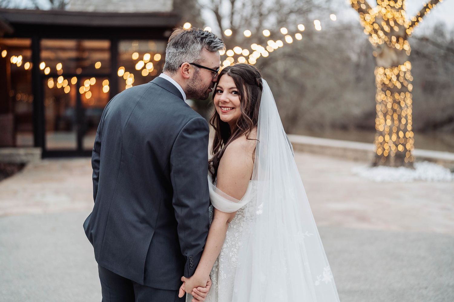 Bride in white gown and veil smiles as groom in gray suit whispers to her outdoors with warm string lights behind.Bride smiling over her sho
