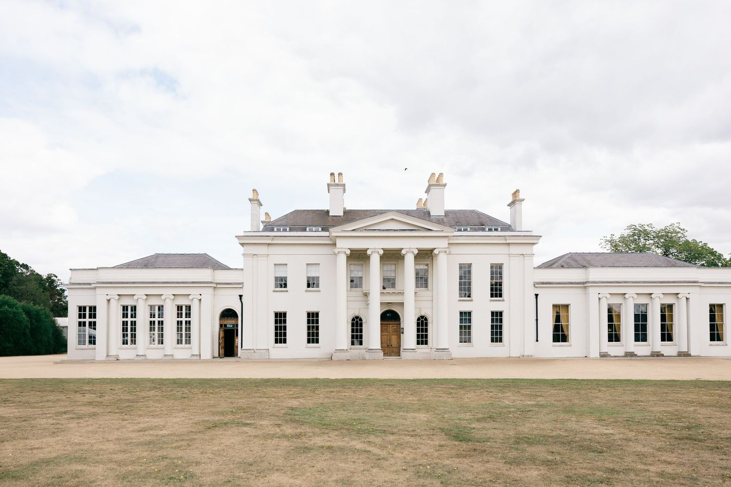 Grand white neoclassical mansion with columned portico set against manicured lawns and cloudy sky.