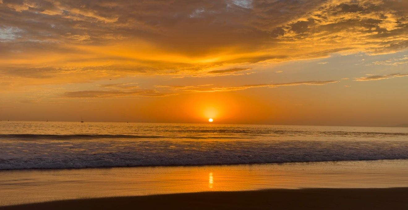 Golden sunset over the ocean with orange clouds, gentle waves, and the sun reflecting on the wet sand of a quiet beach.