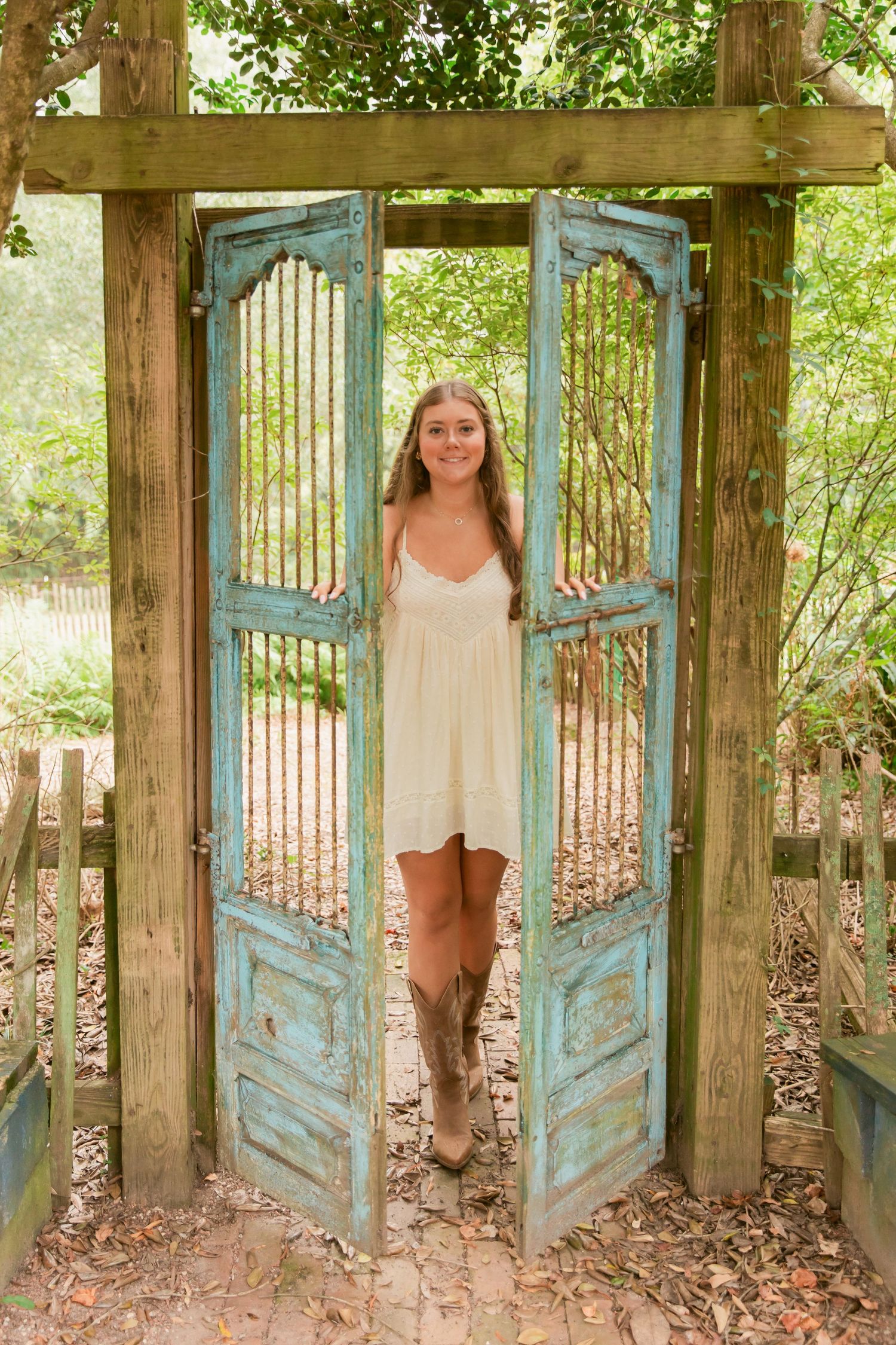 Young woman in white dress and cowboy boots stands in rustic turquoise double doors surrounded by lush greenery.